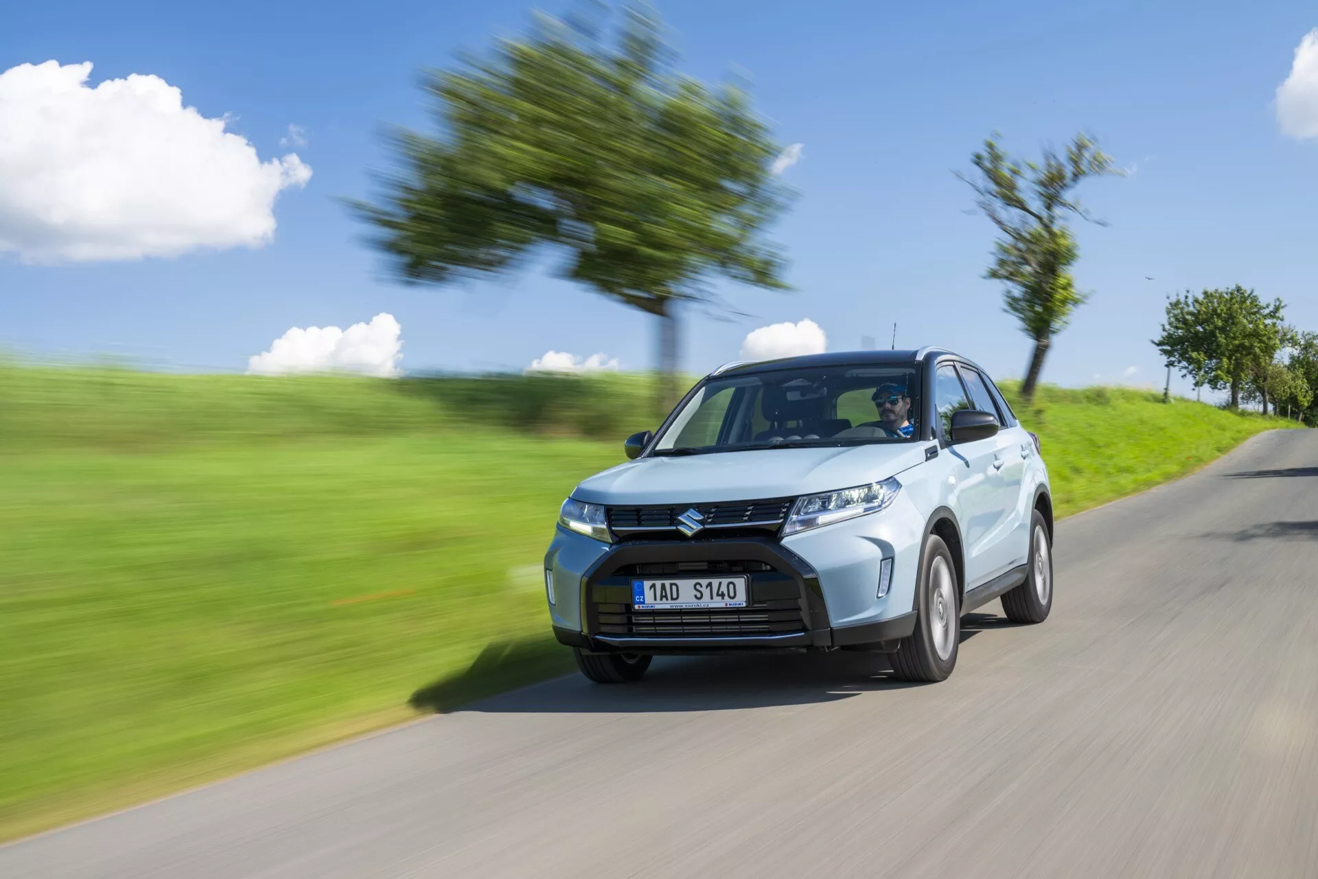 Dynamický snímek Suzuki Vitara 2024 (2. generace facelift), který ukazuje její moderní design a rychlost. Zachyceno z pohledu přední levé čtvrti, obraz zdůrazňuje ostré světlomety, nápadnou přední mřížku a standardní slitiny kol. Efekt rozmazání pohybu na pozadí a kolech zvýrazňuje rychlý pohyb auta, jak je vidět na rozmazané silnici s rostlinami a stromy.