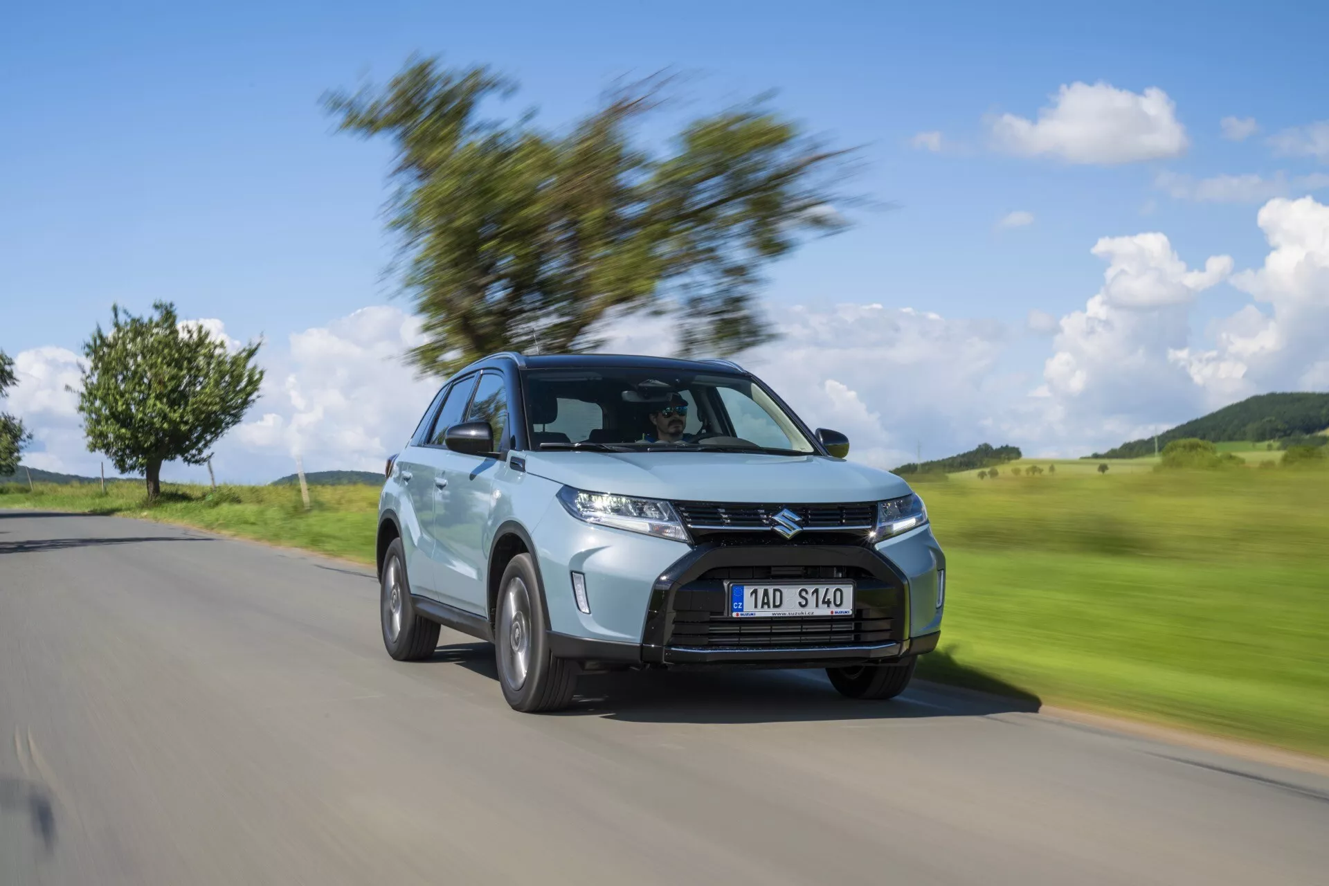 Dynamický obrázek Suzuki Vitara 2024 (2. generace, facelift 2) v pohybu, zachycující přední tříčtvrteční úhel. Auto je ostře zaostřeno, zatímco pozadí je rozmazané pohybem, vyzdvihující jeho světlomety, výraznou masku chladiče, kola z lehkých slitin, karoserii kompaktního SUV, metalickou barvu a aerodynamický design. Ideální pro automobilovou prezentaci a propagační účely.