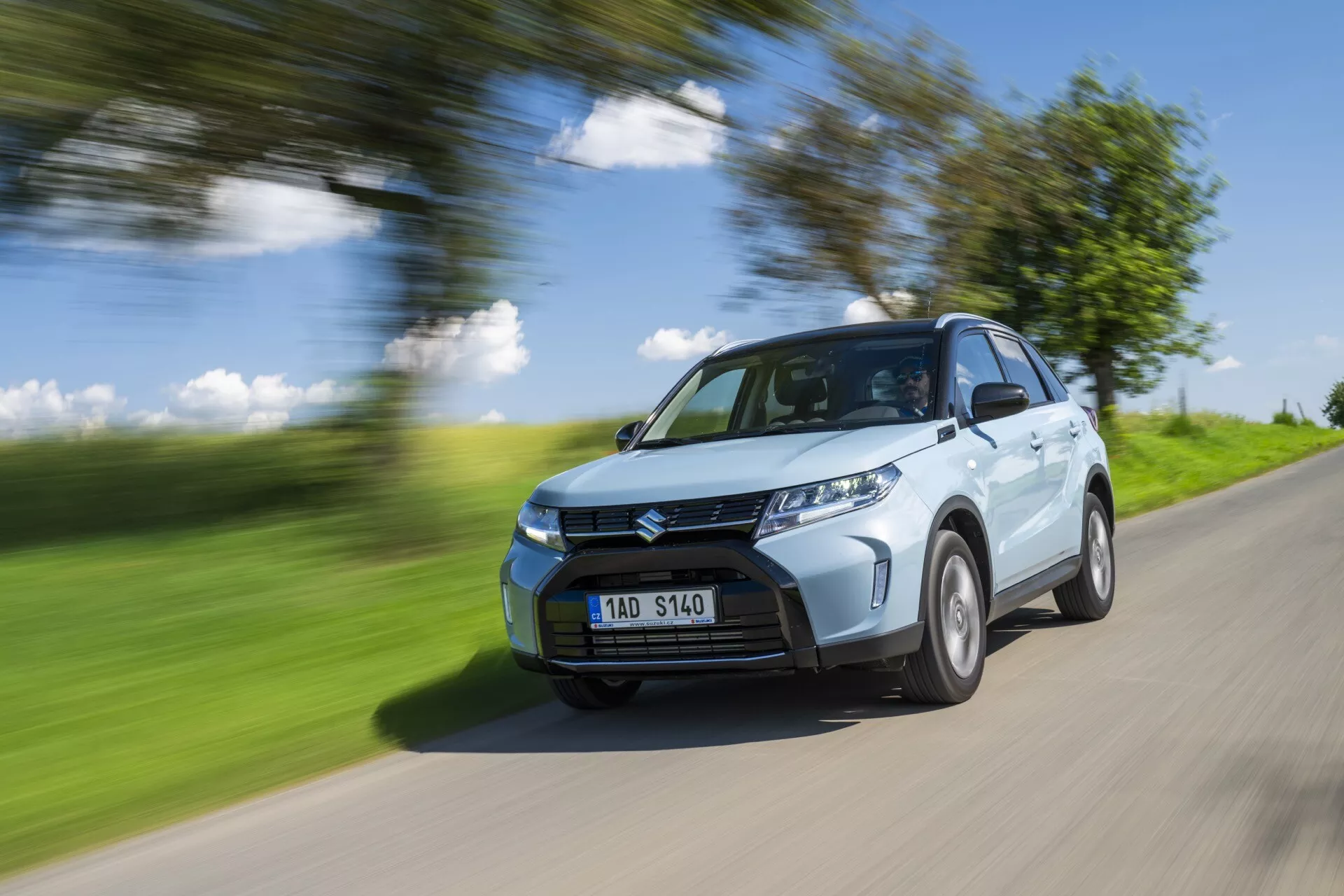Modrá kompaktní SUV Suzuki Vitara 2024 (2. generace facelift) v pohybu na venkovské silnici. Obrázek zachycuje přední čtvrtinu vozu, ukazuje přední masku, světlomety, část kapoty, čelní sklo a stranu řidiče včetně blatníku předního kola, části zadních dveří a části oblouku zadního kola. Auto je ostré oproti rozmazanému pozadí a silnici, což naznačuje rychlost. Kola jsou mírně rozmazaná, což naznačuje rotaci. Osvětleno jasným přirozeným světlem ve slunečný den, s vrhajícími stíny na spodní části vozu, snímek zvýrazňuje designové prvky, jako jsou záhyby na kapotě, design předního nárazníku a vzor mřížky. Světlomety LED jsou patrné, což může naznačovat moderní funkce, jako jsou parkovací senzory. SPZ je viditelná bez citlivých informací.