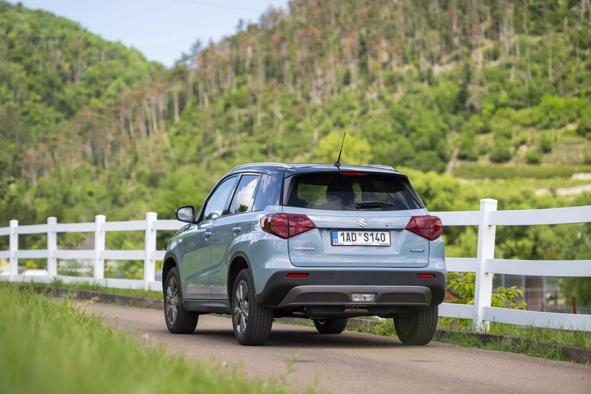 Obrázek ukazuje Suzuki Vitara 2024 (2. generace, facelift) z úhlu tří čtvrtí zezadu, zdůrazňující profil zadní pravé strany při postavení na silnici a mírném naklonění doleva. Viditelné prvky zahrnují zadní dveře, zadní okno, částečné dveře a okna na pravé straně pro pasažéry, LED zadní světla, parkovací senzory na zadním nárazníku, střešní spoiler a krátkou střešní anténu. Auto má litá kola a viditelnou registrační značku. Je zasazeno do venkovského prostředí s bílým plotem a bujnou zelení.