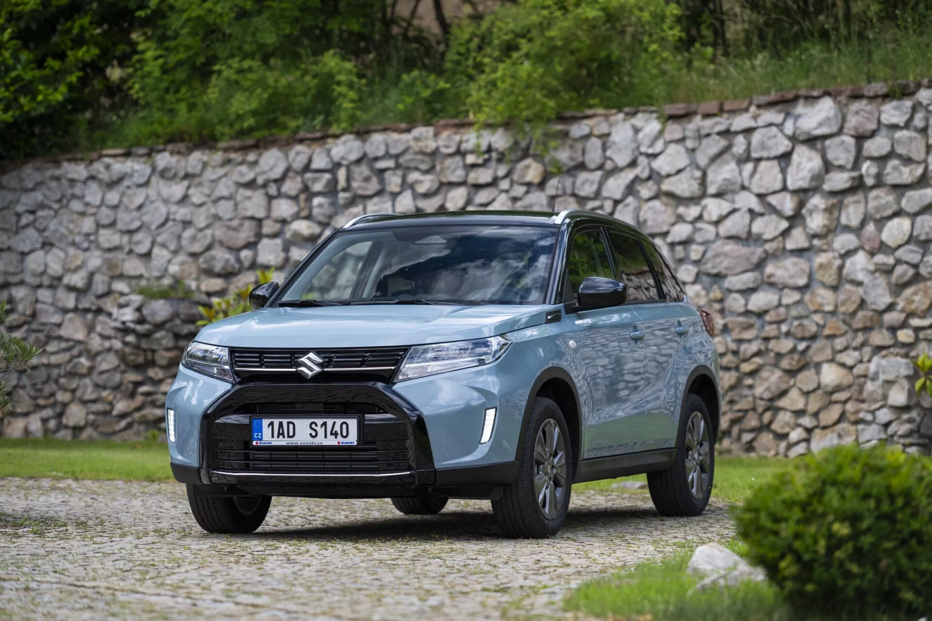 Vysoce kvalitní fotografie automobilu Suzuki v exteriéru, zachycena z úhlu tři čtvrtiny přední strany blízko zeleně nebo parku. Pohled zahrnuje přední masku s výrazným odznakem Suzuki, moderně vypadající světlomety pravděpodobně s LED technologií, mlhové světla v nárazníku a profil řidičovy strany s viditelnými koly, dveřmi a zrcátky. Auto stojí na štěrkovém povrchu s kamennou zdí v pozadí, zdůrazňující přírodní, rustikální atmosféru. Měkce osvětlená, zamračená scéna má mělkou hloubku ostrosti, rozmazávající pozadí, aby se zaměřila na design a technologické detaily auta.