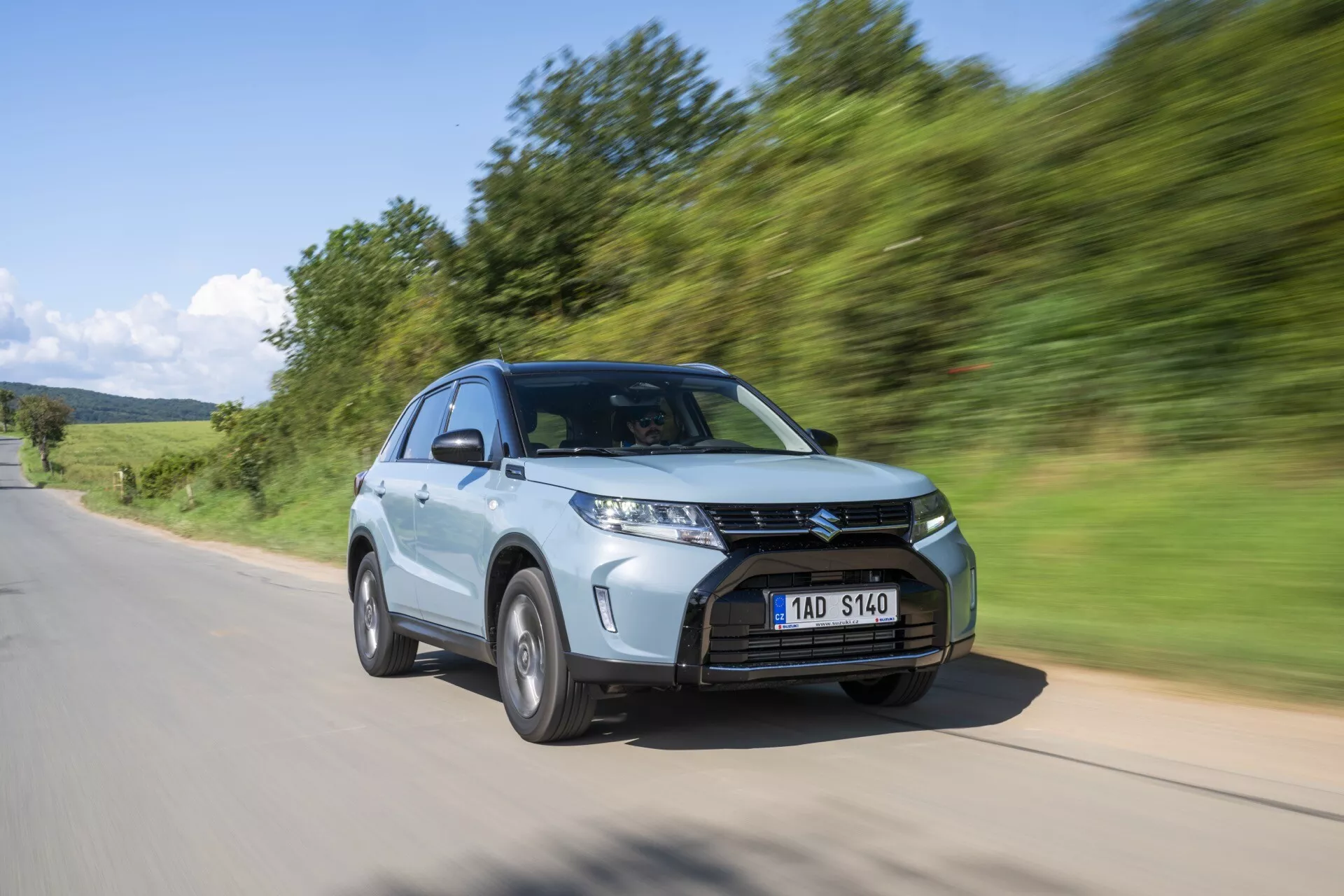 Moderní SUV, 2024 Suzuki Vitara (2. generace, facelift 2) v pohybu z nízkého a tříčtvrtečního úhlu. Obrázek zdůrazňuje výraznou přední část vozidla včetně výrazně navržené masky chladiče s horizontálními lamelami, ostrých a úhlových LED světlometů a multifunkčního hliníkového kola. SUV je zachyceno na asfaltové cestě s přirozenou zelenou krajinou v pozadí, s rozmazáním pohybu, které vyjadřuje rychlost a dynamiku. Další vlastnosti zahrnují šikmou kapotu, aerodynamická zrcátka pro lepší aerodynamiku, vyšší výšku od podvozku a prostorný interiér, vše zvýrazněné metalickým lakem.
