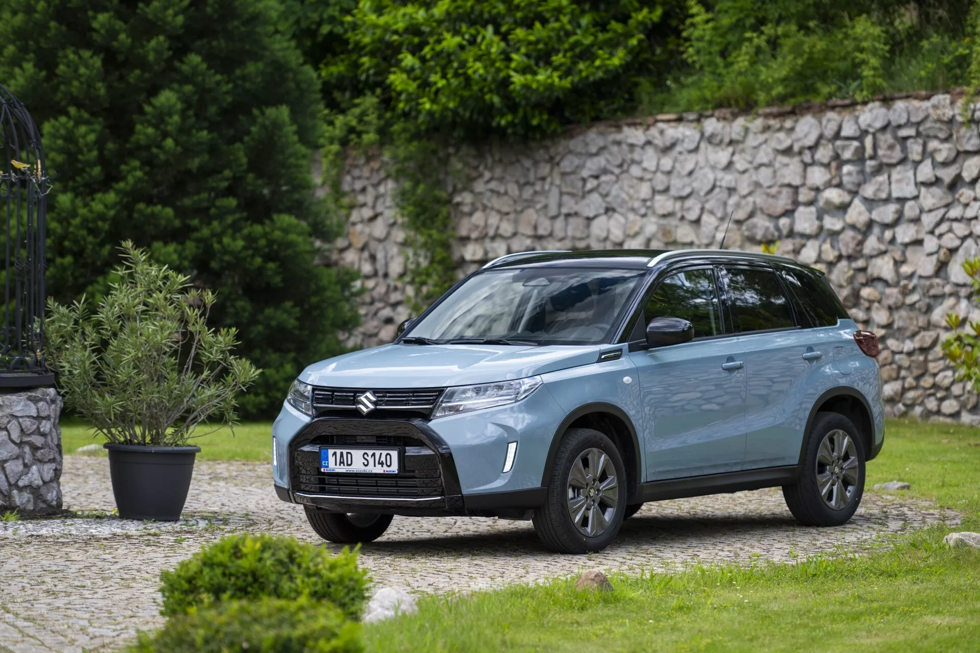 Šikmý přední boční pohled na moderní SUV Suzuki Vitara 2024 zaparkované venku v rezidenční nebo parkové oblasti s dobře udržovaným trávníkem a kamenou zídkou v pozadí. Obraz, pořízený ze střední vzdálenosti, ukazuje přední mřížku s horizontálními pruhy s logem společnosti, elegantní LED světlomety s denními světly, sochou tvarovaný nárazník s integrovanými mlhovkami, hliníková kola, integrovaná směrová světla v bočních zrcátkách, kovový povrch laku, dynamické linie karoserie, tónovaná okna, střešní nosiče a anténu na střeše, podtrhující jeho moderní design a vlastnosti.