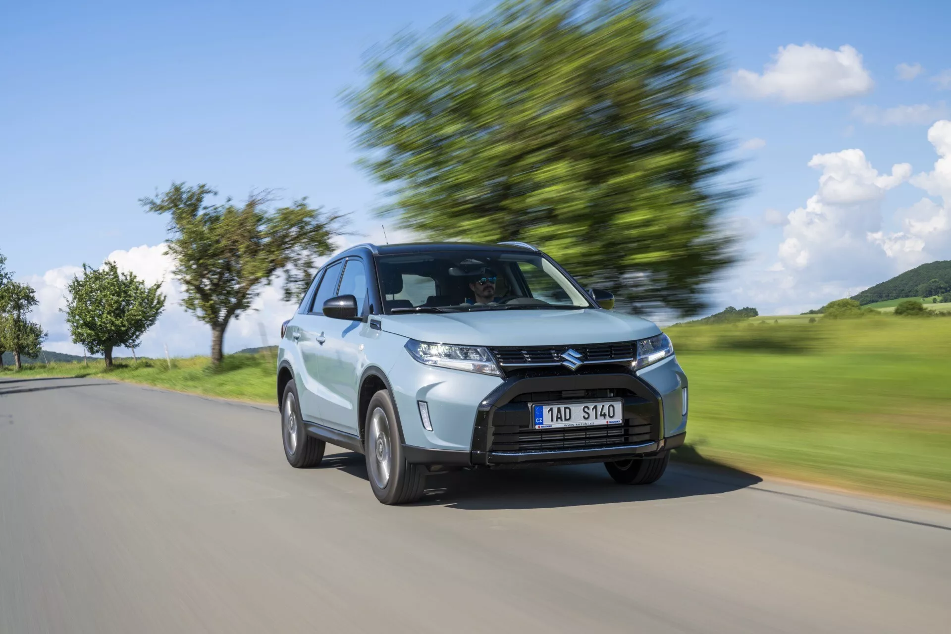 Dynamický obraz stříbrného hatchbacku Suzuki Vitara 2024 v pohybu zachycený z tři čtvrtinové přední strany, který zdůrazňuje moderní design s výraznou maskou, hranatými světlomety, střešní linií a koly v pohybu s rozmazaným pozadím znázorňujícím rychlost.