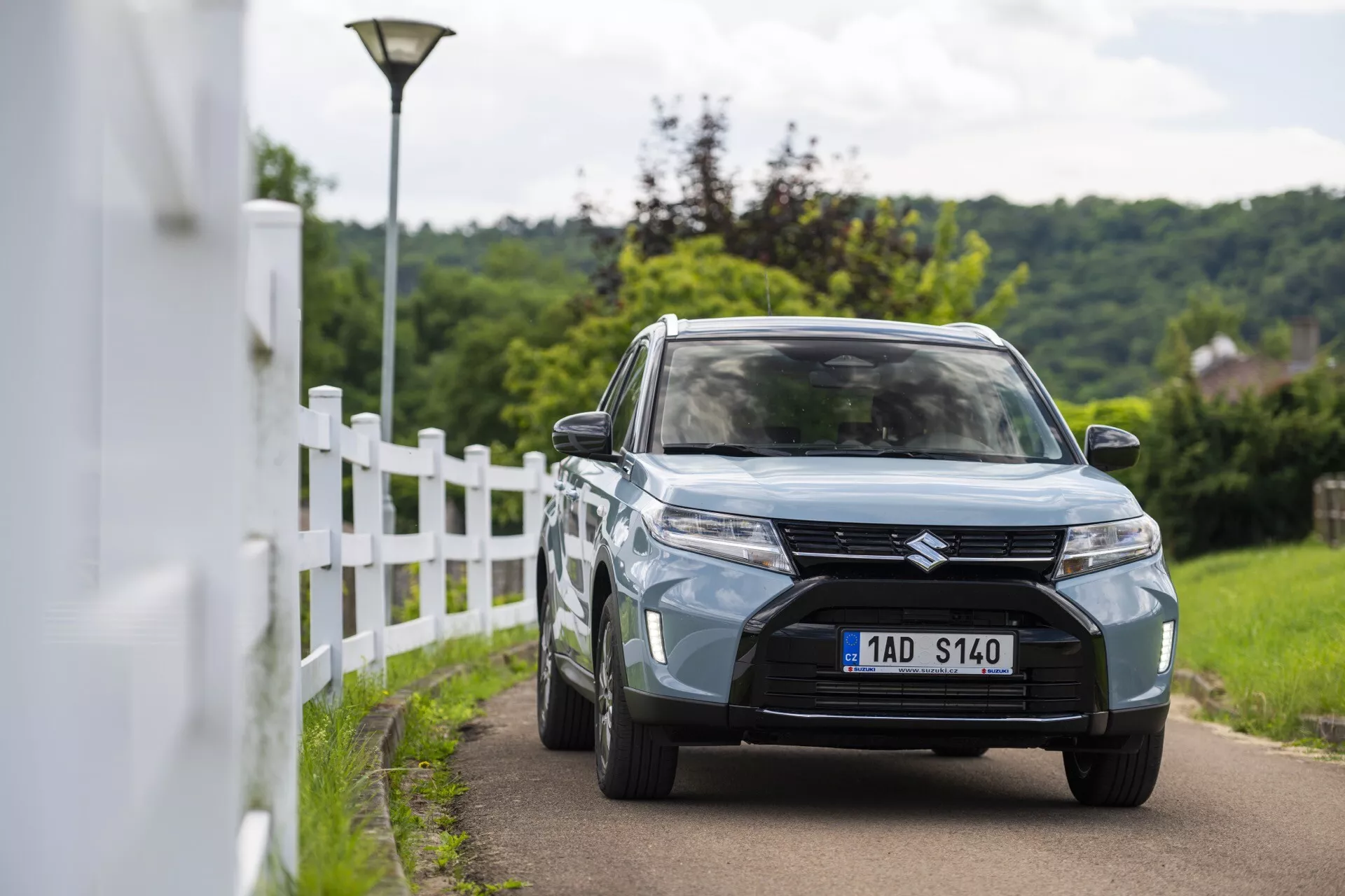 Denní scéna venku zobrazující Suzuki Vitara 2024 (2. generace facelift) zachycený z předního tříčtvrtečního pohledu. Obrázek zdůrazňuje designové prvky vozidla včetně výrazné přední masky s horizontálním šprušlovým designem a centrálním odznakem, moderní LED nebo HID světlomety, zapuštěné mlhovky ve spodním nárazníku a světle modré nebo šedé barvy. Auto jede na úzké silnici poblíž bílého plotu s mírně rozmazaným pozadím, což naznačuje malou hloubku ostrosti. Viditelné prvky zahrnují také slitinová kola, boční zrcátka s integrovanými směrovkami a částečně viditelný přední nárazník s centrálním vzduchovým přívodem.