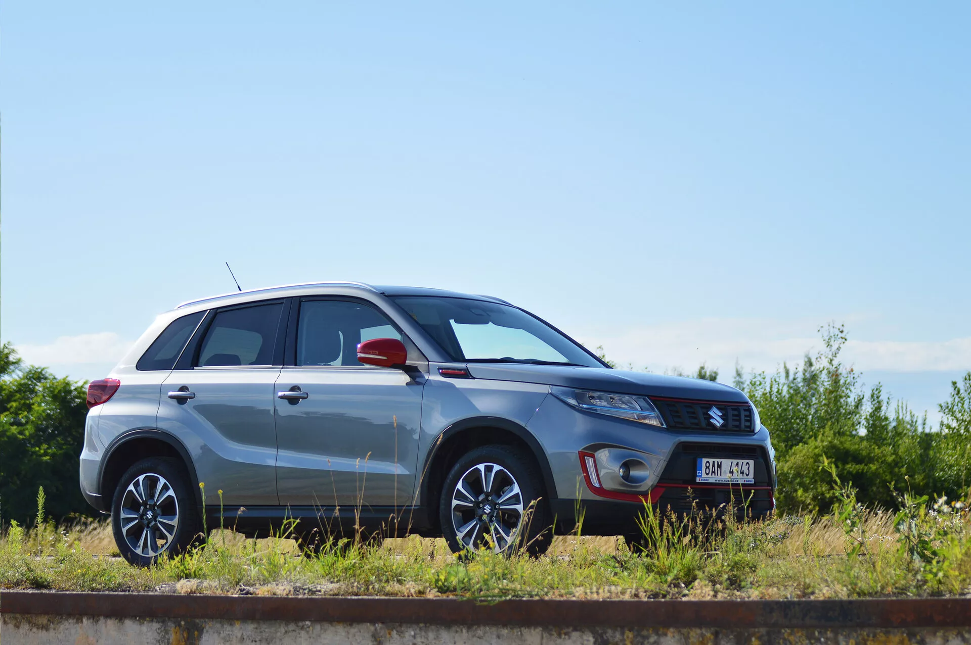 Boční profil stříbrného kompaktního SUV Suzuki Vitara 2018, zaparkovaného paralelně s železniční tratí za slunečného dne. Jsou zřejmé designové linie, kola z lehkých slitin s vícepaprskovým designem, metalický lak, zavřené dveře a okna, tvar světlometů, boční zrcátko s potenciálními integrovanými směrovkami a označení výbavy, což ukazuje design a technologické prvky vozu.