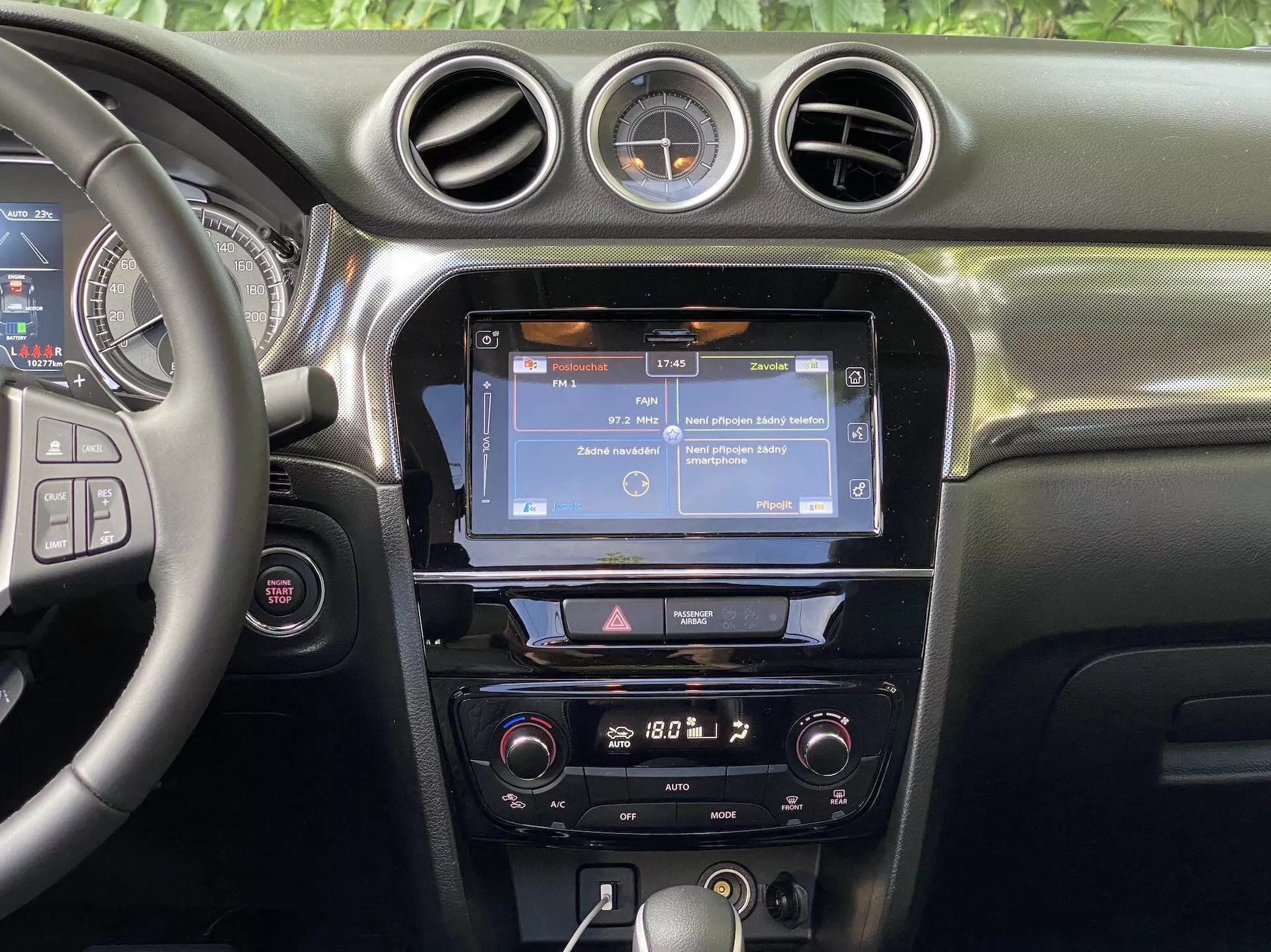 Pohled na interiér modelu Suzuki Vitara 2018 se zaměřením na středovou palubní desku. Sekce obsahuje částečně viditelný volant vlevo s několika ovládacími tlačítky, pravděpodobně pro ovládání audia, telefonování hands-free a tempomatu. Středová konzole zobrazuje dotykovou obrazovku infotainmentu, pravděpodobně zobrazující navigaci nebo menu, obklopenou vertikálními ventilačními otvory a doplněnou třemi kruhovými otvory. Pod dotykovou obrazovkou je řada tlačítek a ovládacích prvků pro rychlý přístup k nastavením infotainmentu a vozidla. Ovládání klimatizace je umístěno dále dole, což naznačuje dvouzónový klimatizační systém. Palubní deska je vybavena měkkým povrchem se viditelným prošíváním a mezi centrálními kruhovými ventilačními otvory je umístěn kulatý analogový hodiny. Viditelnost je zaměřena na prvky palubní desky, bez oken nebo čelního skla v záběru.