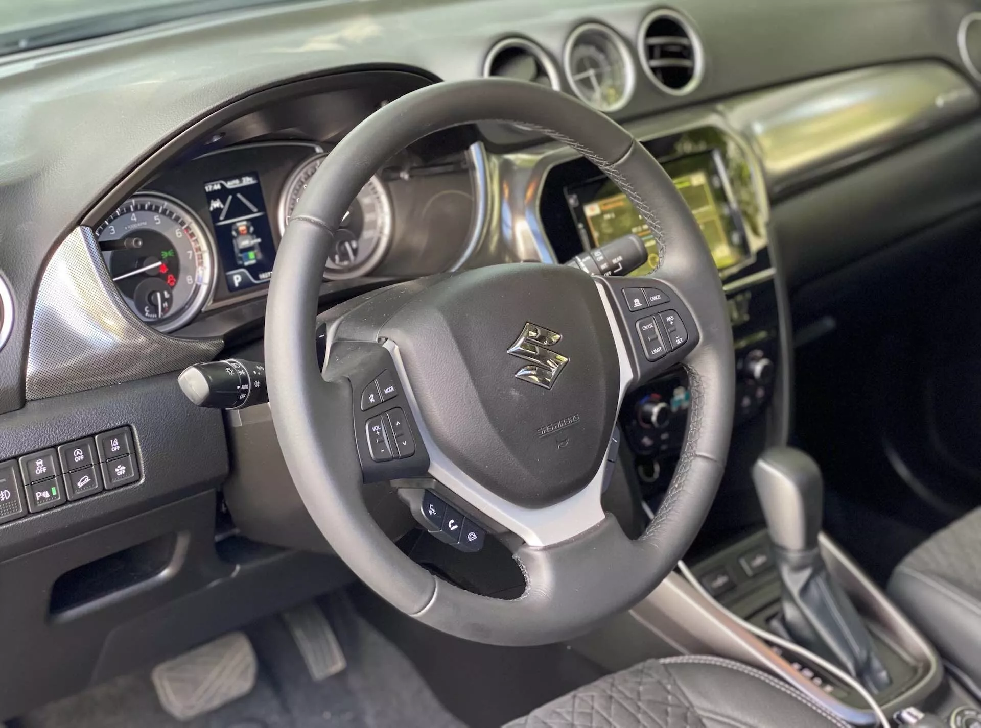 Interiér Suzuki Vitara 2018, z perspektivy řidiče. Detail koženého vícefunkčního volantu s emblémem, ovládacími prvky pro zvuk, hlasové ovládání a tempomat s kovovými akcenty. Za ním je přístrojová deska s analogovými číselníky pro rychloměr a otáčkoměr a digitální obrazovkou pro informace o vozidle, indikující, že auto je vypnuté nebo v režimu příslušenství. Středová konzole ukazuje kovovou nebo chromovou hromádku s dotykovým infotainment systémem, tlačítky a knoflíky pro ovládání klimatu a vozidla. Automatická řadící páka s koženými akcenty naznačuje různé režimy jízdy. Interiér kombinuje kůži a kartáčovaný kov, což odráží moderní design. Předpokládané pokročilé funkce zahrnují Bluetooth, navigaci, parkovací asistenty a systémy asistence řidiče.