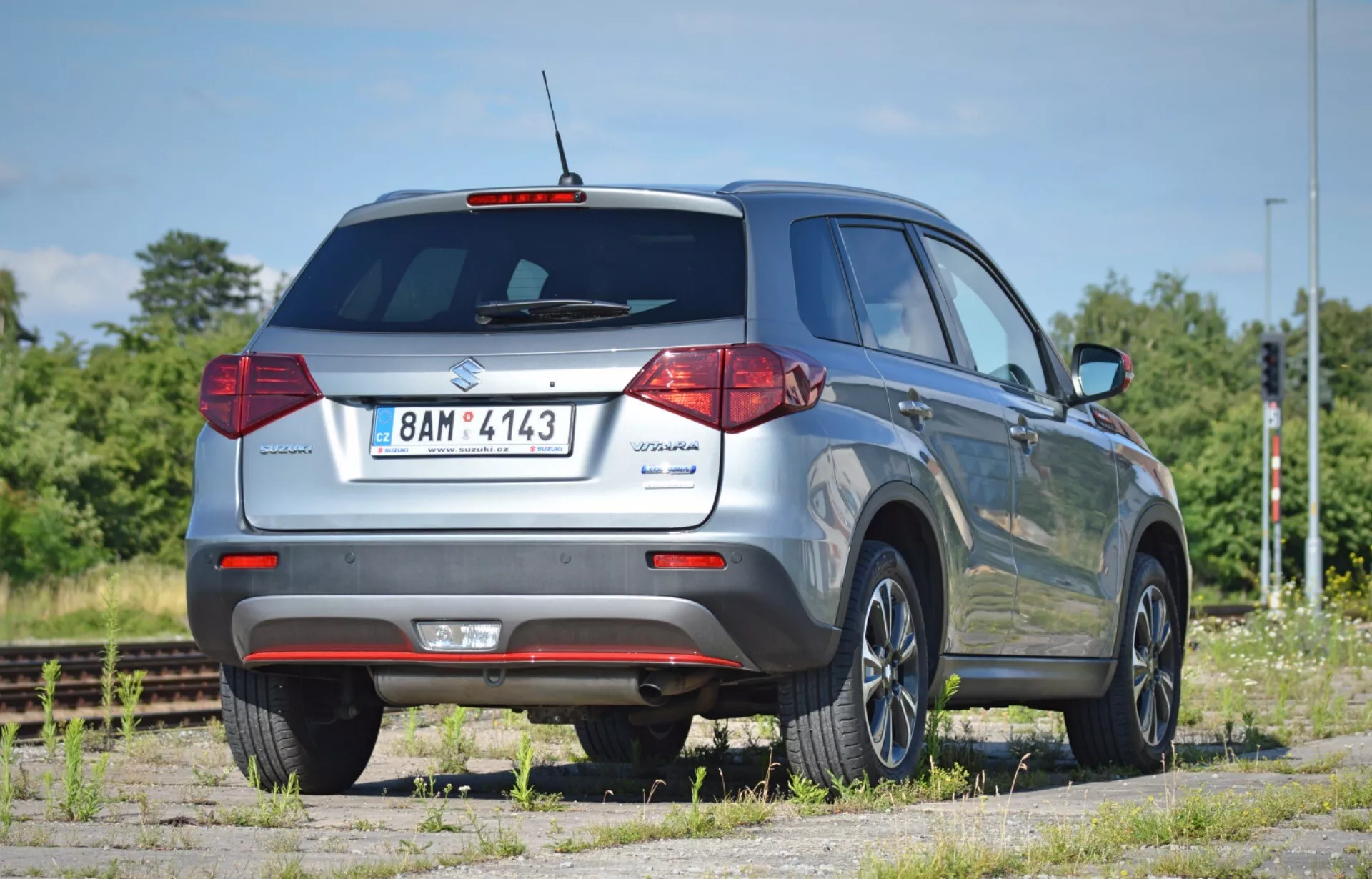 Zadní pohled na SUV Suzuki Vitara 2018 z nízkého úhlu, zvýrazňující jeho výšku a průmět světlé výšky. Vozidlo je umístěno pod úhlem 45 stupňů, což ukazuje zadní dveře s odznakem modelu, zadní okno se stěračem, tradiční halogenová zadní světla, reflexní zadní nárazník a viditelnou výfukovou trubku. Pravá strana zahrnuje zadní dveře pro pasažéry, boční okna, střešní ližinu a standardní litá kola. Na bočních zrcátkách nejsou viditelné žádné ukazatele směru. Vozidlo je zaparkované na prašném povrchu poblíž železničních kolejí, v suché, slunné oblasti se zelení a modrou oblohou.