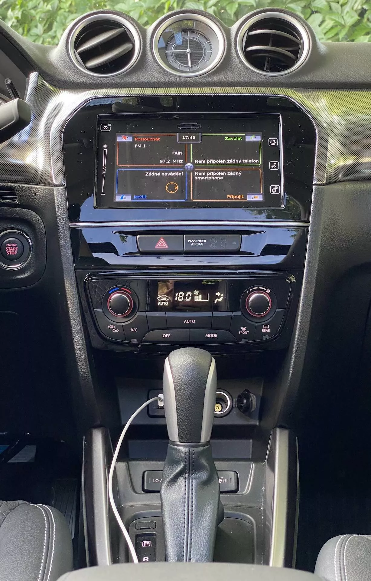 Podrobný pohled na interiér Suzuki Vitara 2018, zdůrazňující středovou konzoli a palubní desku z pohledu předních sedadel. Obrázek zdůrazňuje dotykovou obrazovku infotainmentu zobrazující nabídku, dvouzónový klimatizační systém s digitálními ukazateli a automatickou převodovku. Interiér ukazuje tmavé barvy s mixem matných a lesklých textur, ilustrující kvalitu materiálu a moderní automobilovou technologii.