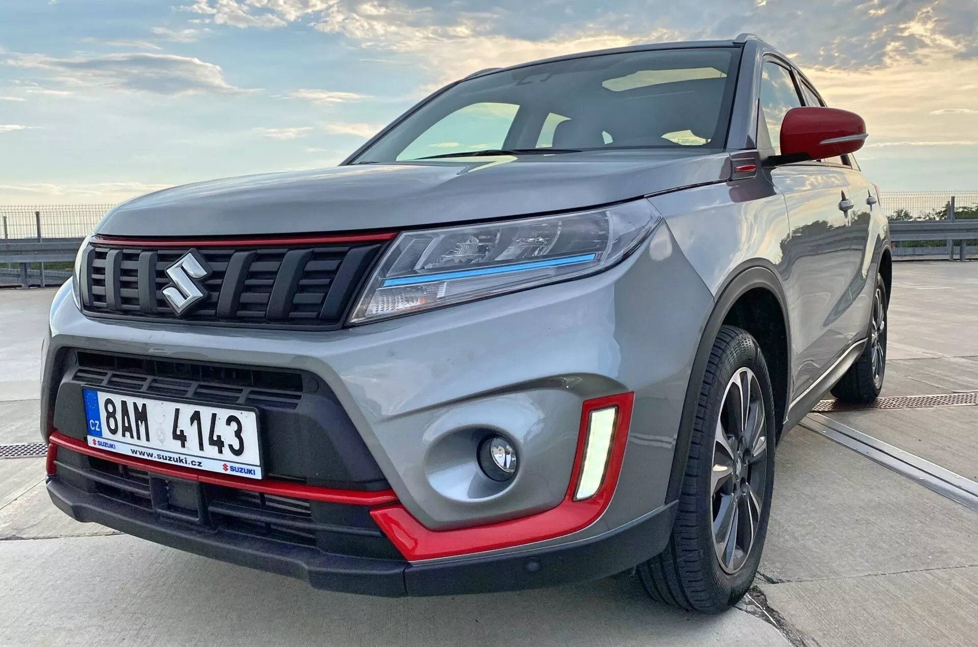 Blízký pohled na moderní kompaktní SUV z boční přední strany, šikmo ukazující část bočního profilu. Obrázek zdůrazňuje robustnost vozu s viditelnými detaily jako přední nárazník, mřížka chladiče s horizontálními lamelami a emblémem, elegantní LED světlomety, mlhové světlomety s červenými vložkami, litá kola a svalnaté blatníky. Tmavá litá kola a kontrastní barevné prvky naznačují prémiovou nebo sportovní výbavu.