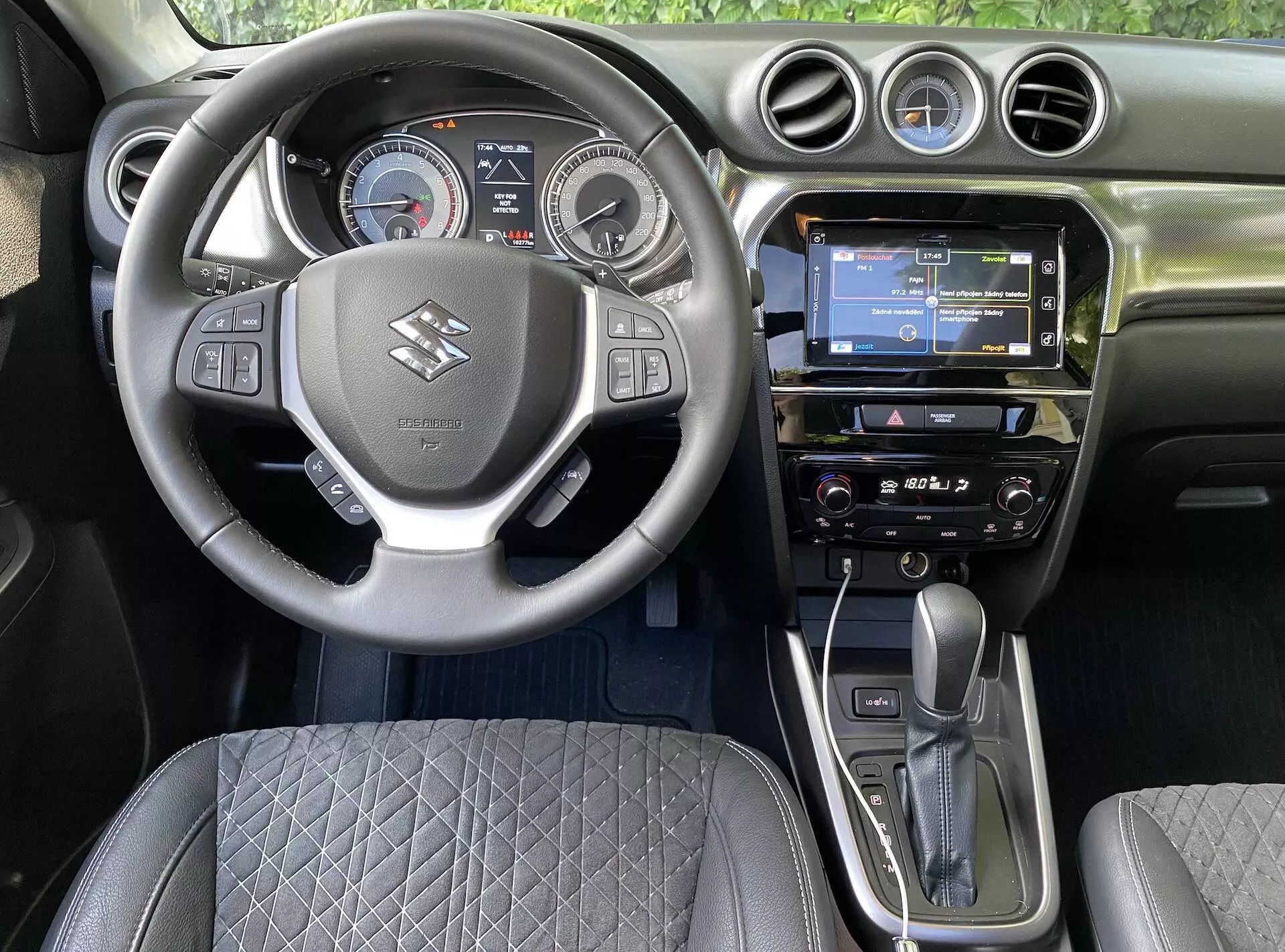 Pohled na interiér Suzuki Vitara 2018 z místa řidiče, ukazující středový volant s logem značky, ovládacími tlačítky pro hlasitost, hlasové příkazy a připojení telefonu. Za volantem je digitálně-analogová přístrojová deska s rychloměrem, otáčkoměrem a centrálním digitálním displejem. Palubní deska obsahuje dotykovou infotainment obrazovku s GPS navigací, rádio a klimatizaci. Automatická řadicí páka a elektronická ruční brzda jsou výrazně zobrazeny, spolu s prošívaným designem sedadla a kruhovými větracími otvory, zachycující moderní automobilovou technologii a ergonomii designu.