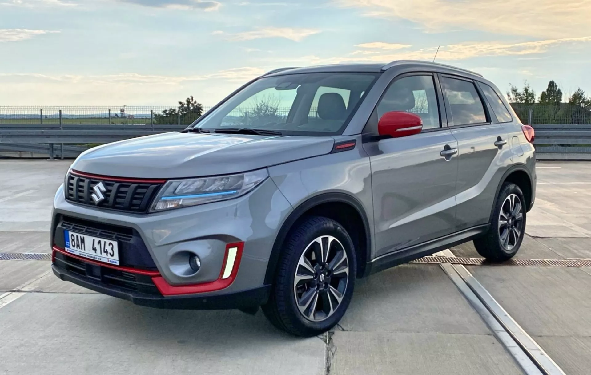 Obrázek ukazuje kompaktní SUV Suzuki Vitara z roku 2018 zaparkované v exteriéru na parkovišti během východu nebo západu slunce. Fotografie zachycuje vůz z tříčtvrtečního předního úhlu a odhaluje tak řidičovu stranu a předek, zvýrazňujíc konfiguraci světlometů, styl mřížky, přední nárazník, kola a boční linie karoserie. Vozidlo má dvoubarevný nátěr s hlavním tělem ve stříbřitě šedé barvě a černou střechou a zpětnými zrcátky, doplněné červenými akcenty na předním nárazníku. Viditelné jsou také elegantní LED světlomety, výrazný znak značky na mřížce se vzorem a vícepaprsková litá kola, což ukazuje moderní a sportovní design vozidla.