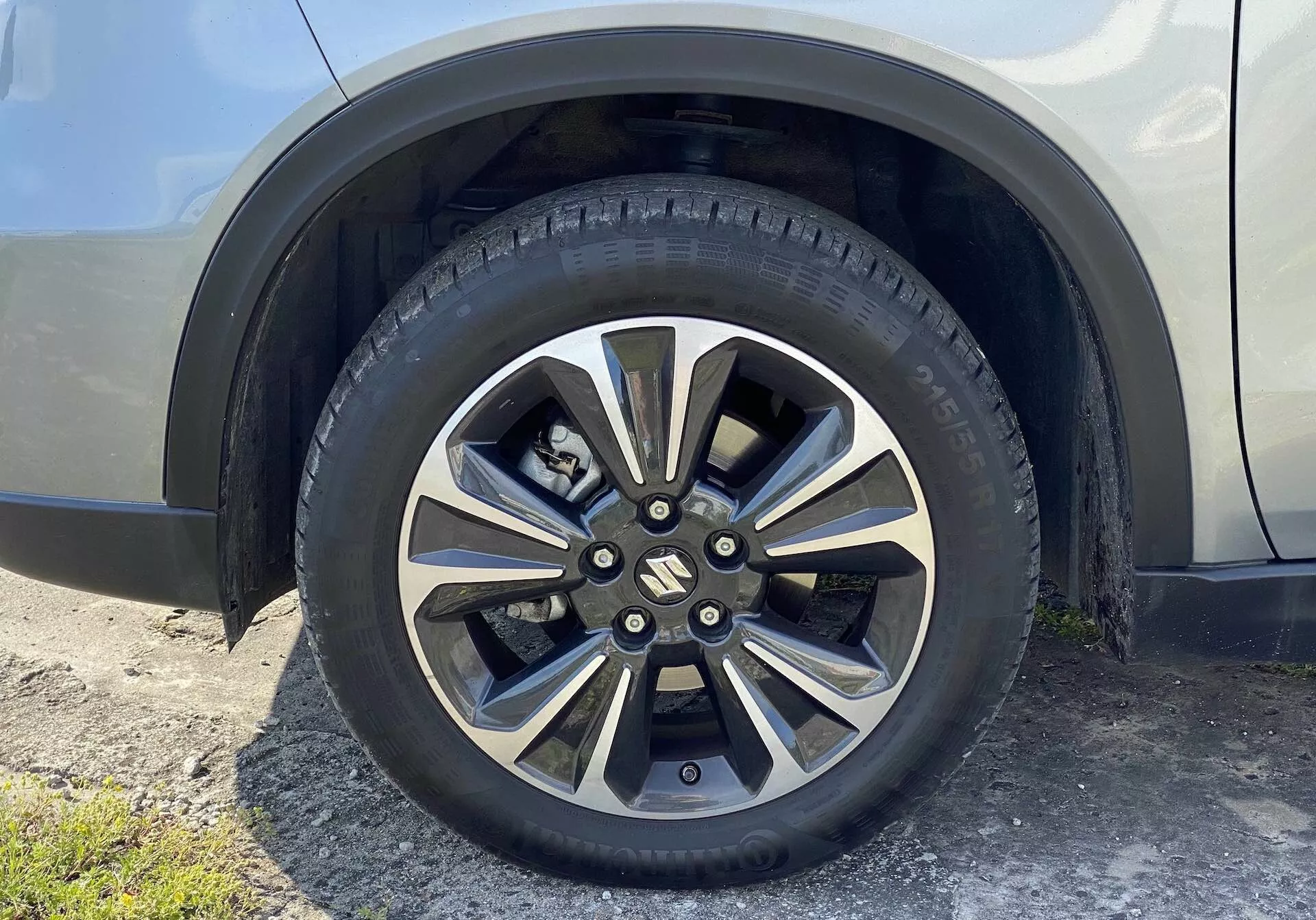 Detailní pohled na kolo Suzuki Vitara 2018, které představuje slitinovou konstrukci s více západkami a metalickými i tmavšími lakovanými částmi. Značený středový kryt zakrývá matice kol a pneumatika určená pro silniční použití obsahuje podrobné vzorky běhounu a označení boční stěny. Pozadí zahrnuje lesklou část blatníku a bočního panelu auta v neutrální barvě, pohled mírně šikmo vzhůru ze země.