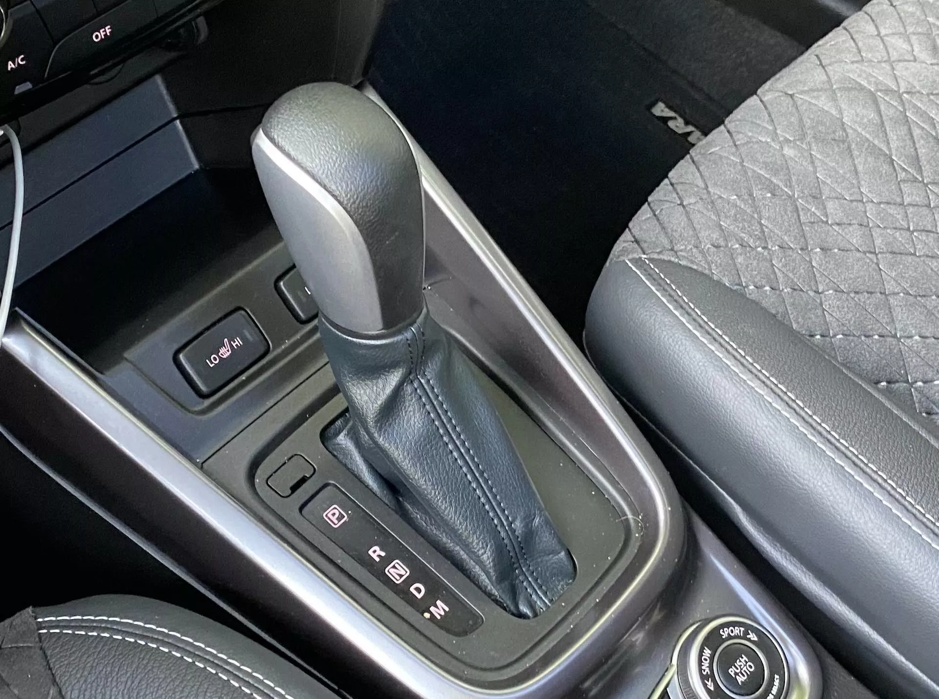 Detailní záběr na středovou konzoli vozu Suzuki Vitara 2018, zaměřující se na řadicí páku v parkovací poloze 'P', obklopenou kovovým pouzdrem s jasnými indikátory rychlostí. Vlevo je vidět ovládací panel se dvěma tlačítky a okraj obrazovky. Středová konzole je vyrobena z vysoce kvalitních materiálů a napravo je patrně část předního sedadla potaženého prošívaným designem. K dispozici je také otočný ovladač pro infotaimentové funkce, což podtrhuje pokročilé technologie a luxusní prvky vozu.