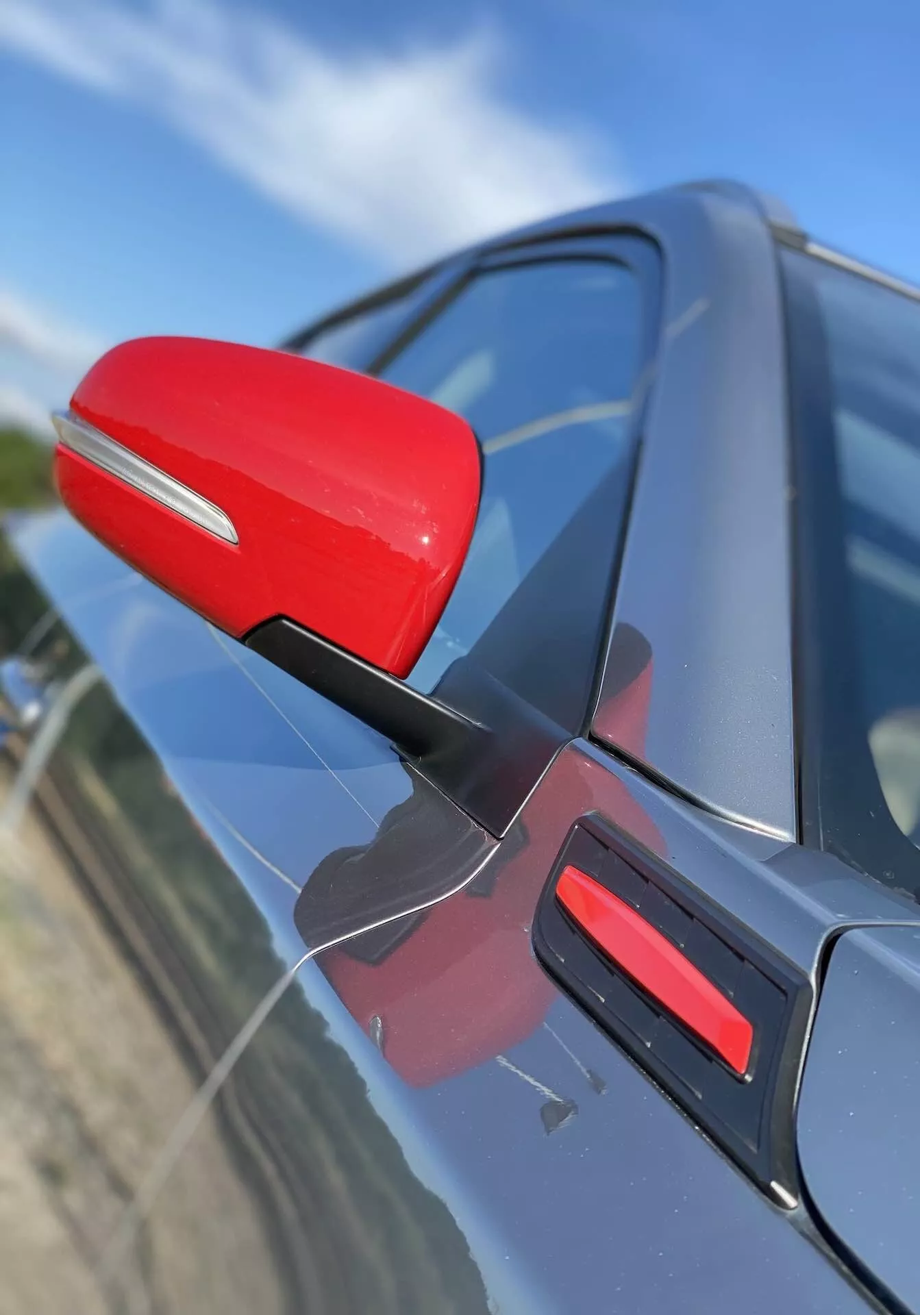 Detailní pohled na zpětné zrcátko na straně řidiče Suzuki Vitara 2018, zvýrazněné červenou barvou proti tmavé barvě karoserie vozu. Zrcátko je připevněno seštíhlenou černou podpěrou, naznačující aerodynamickou účinnost. Záběr zachycuje zrcátko z mírně předního úhlu šikmo, s pohledem dolů, obsahující odpovídající červený prvek na dveřích. Pozadí je rozmazané, zdůrazňující detaily zrcátka a designu dveří auta.