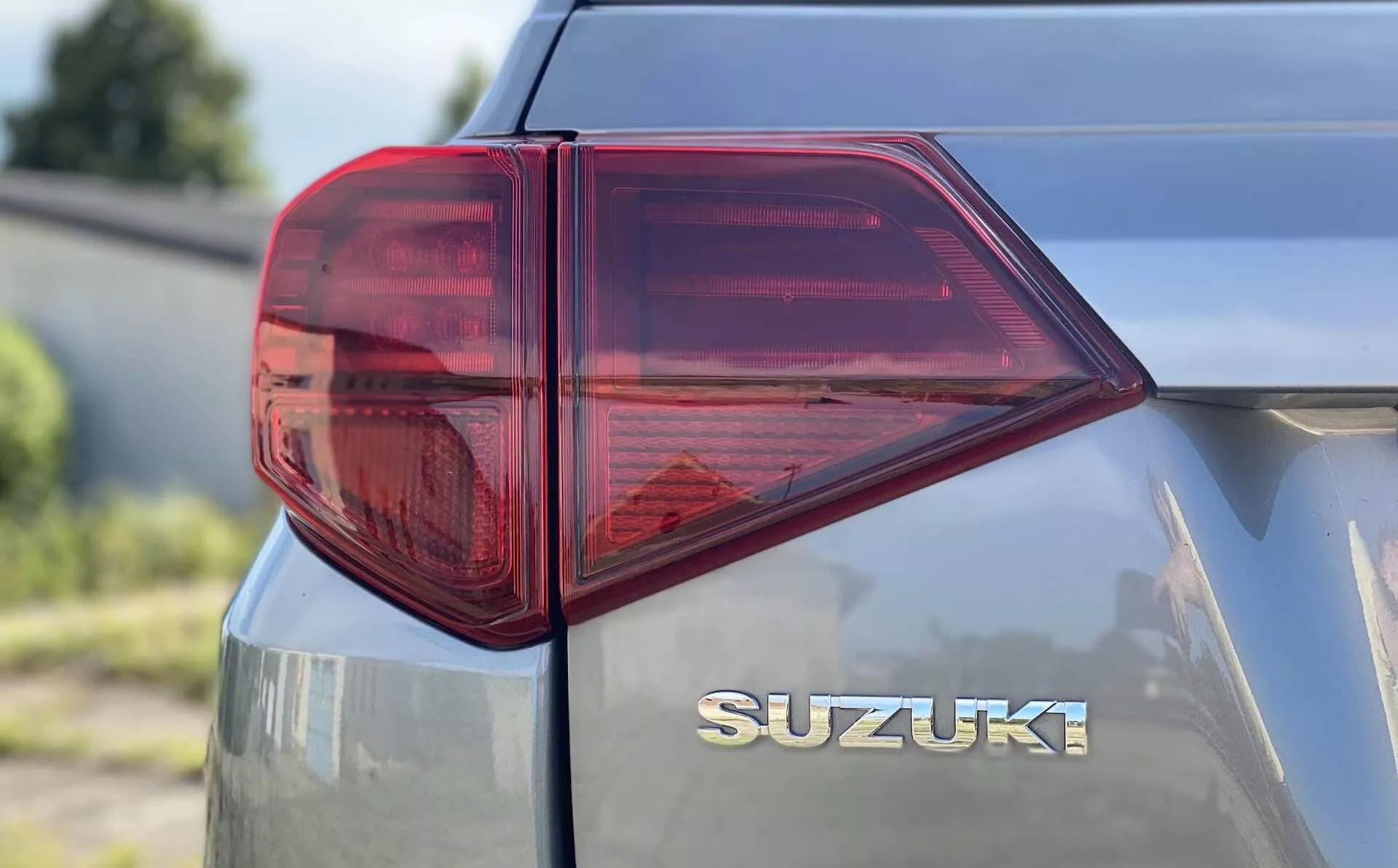 Zblízka z levé zadní strany vozu Suzuki Vitara 2018 zaměřený na zadní světlo. Snímek ve tříčtvrtečním pohledu z boku, pořízený z nízko až středně vysoké výšky, ukazuje výrazně stylizované LED zadní světlo s ostrými úhly a několika komorami pro funkce jako brzdové světlo, směrové světlo a případně mlhovku. Převážně červené světlo s čirou částí, pravděpodobně pro couvací světlo, zdůrazňuje moderní automobilovou osvětlovací technologii. Zadní víko vybavené chromovaným znakem „SUZUKI“ umístěným uprostřed pod zadním oknem zdůrazňuje čisté a hladké značení. Kovový lak odráží trochu světla, což naznačuje dobře udržovaný nebo novější povrch vozidla.