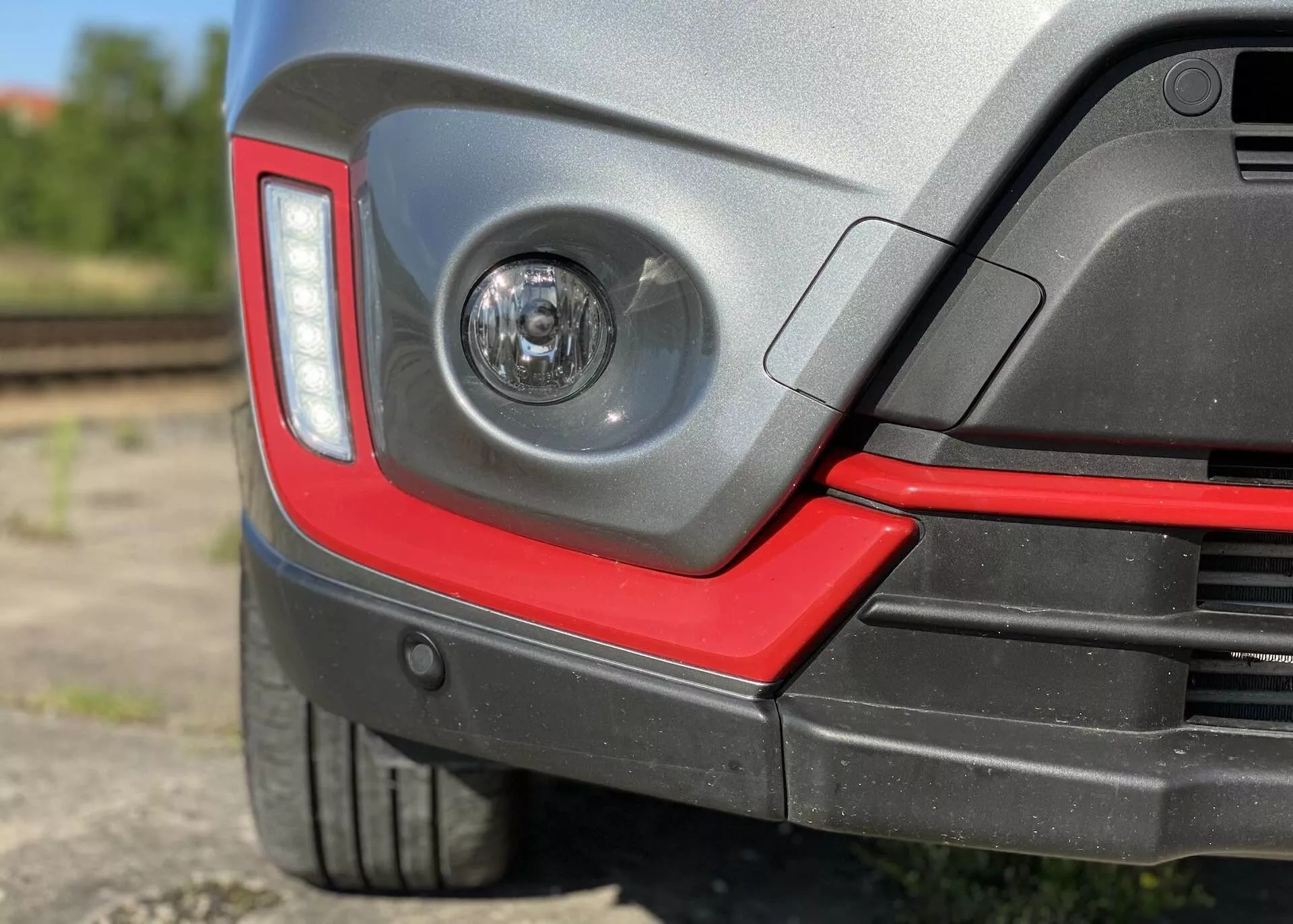 Detailní pohled na zadní roh vozu Suzuki Vitara 2018, který zahrnuje čirý mlhový světlomet v zadním nárazníku, moderní hranatý design zadních světel, dvoubarevný zadní nárazník se sportovním červeným akcentem, potenciální parkovací senzory, nízkoprofilovou pneumatiku s ochranným plastovým obloukem, lesklý lak a rozmazané poloprůmyslové pozadí s železničními kolejemi.