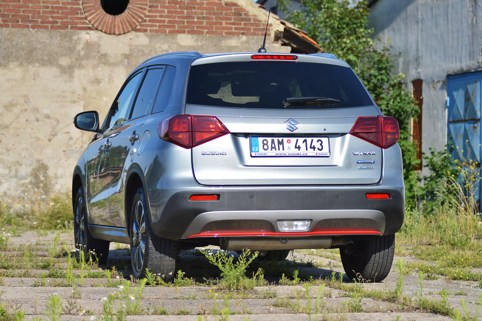 Zadní tříčtvrteční pohled na metalickou šedou Suzuki Vitara 2018 hatchback zachycený za denního světla. Obrázek zdůrazňuje zadní design vozu, včetně výrazných zadních světel, zadního stěrače a emblémů Suzuki a Vitara. Také ukazuje odznak 'Allgrip' označující pohon všech kol, evropskou registrační značku, litá kola, parkovací senzory na zadním nárazníku a anténu na střeše. Vozidlo je zaparkované na nerovnoměrném povrchu s trávou a hlínou, což naznačuje venkovní, možná zanedbanou, oblast. Auto je čisté a dobře udržované.