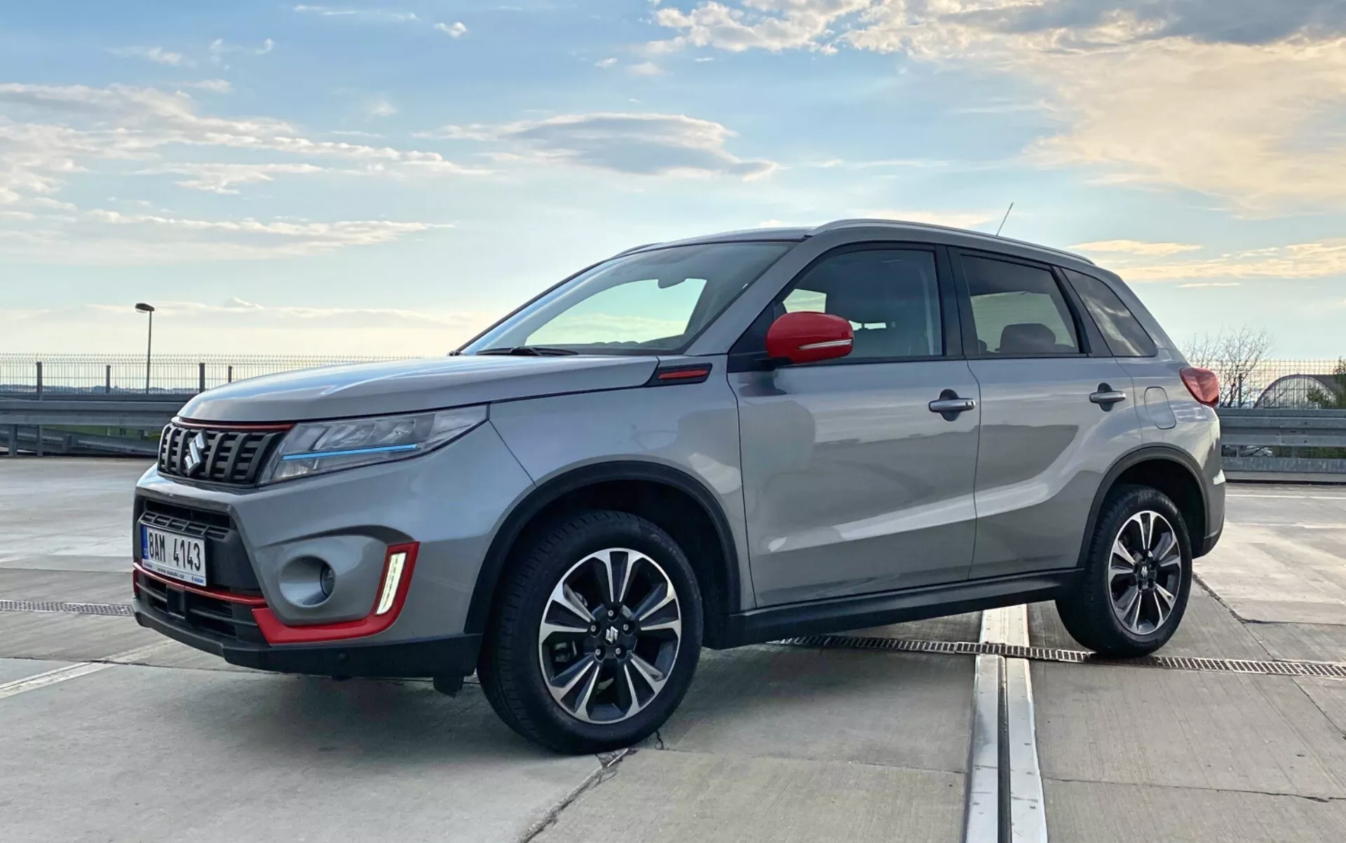 Obrázek zobrazuje Suzuki Vitara 2018, moderní kompaktní SUV, zaparkované venku na denním světle. Je zachyceno z tříčtvrtečního předního pohledu, zdůrazňujícího jak přední, tak boční profil. Úhel je mírně nízký, na úrovni klik dveří, což naznačuje, že fotograf byl v dřepu nebo se ohýbal. Viditelné detaily zahrnují přední mřížku, světla, pouzdro mlhových světel, emblém, boční zrcátka, litá kola s integrovanými směrovými světly, kliky dveří, rámy oken, střechu a střešní ližiny. Projektorová světla a elegantní anténa naznačují pokročilé technologie. Moderní designové prvky SUV naznačují funkce jako LED světla a možná bezklíčový vstupní systém.