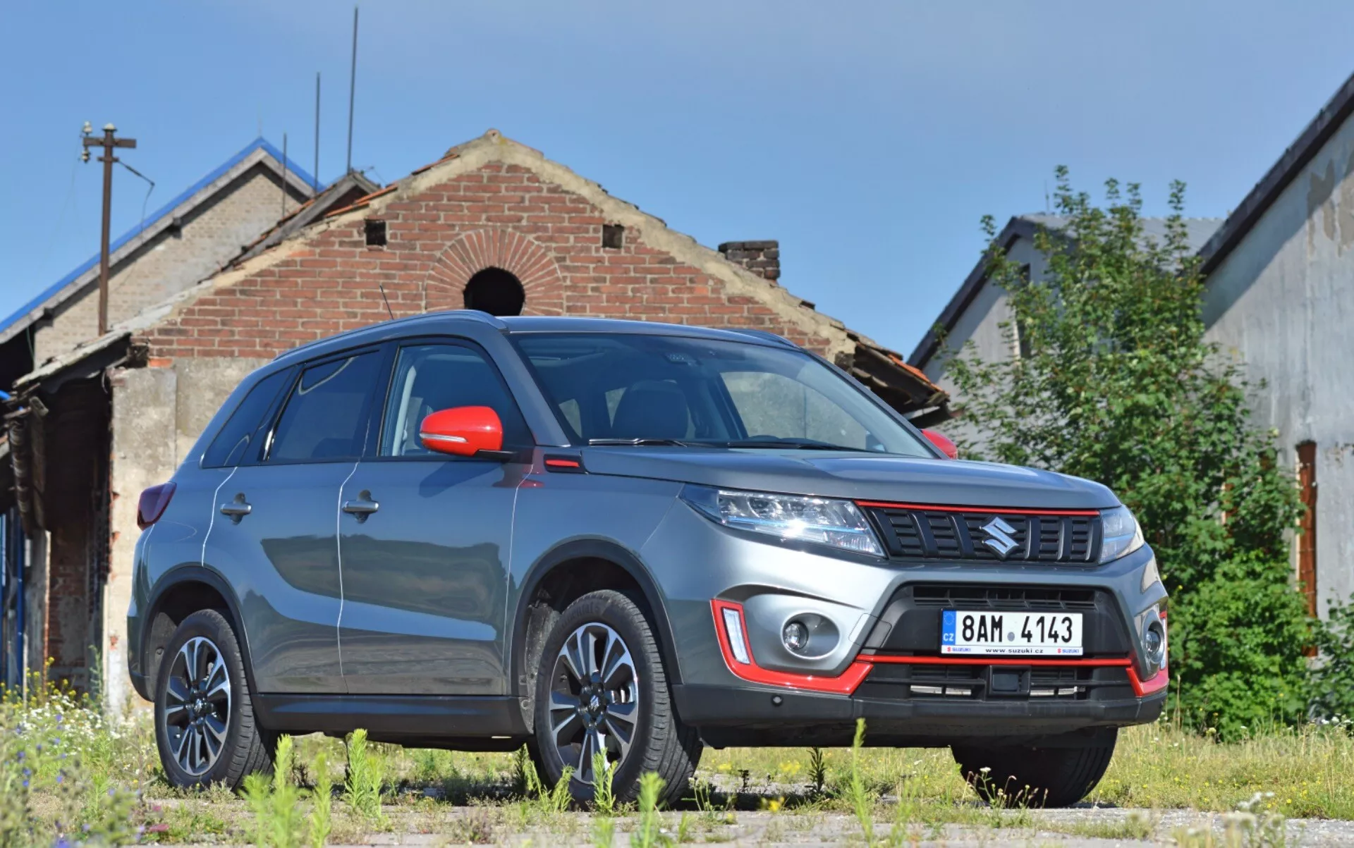 Obrázek představuje SUV Suzuki Vitara 2018 z nízkého tříčtvrtečního předního pohledu, který zvýrazňuje výraznou přední pravou stranu vozidla s velkou světlou výškou. Viditelné jsou robustní přední nárazník s mlhovými světly a stříbrným krytem, emblém značky na horizontální mřížce, výrazné linie kapoty, moderní světlomety možná s LED nebo HID technologií, víceramenná hliníková kola, červená zpětná zrcátka s integrovanými směrovkami, výrazné blatníky pro svalnatý vzhled, reflexní čelní sklo a tónovaná okna. Vitara je zaparkována na nezpevněném travnatém povrchu před starou budovou, což dodává průmyslový nebo venkovský kontext, což naznačuje vhodnost pro venkovní prostředí.