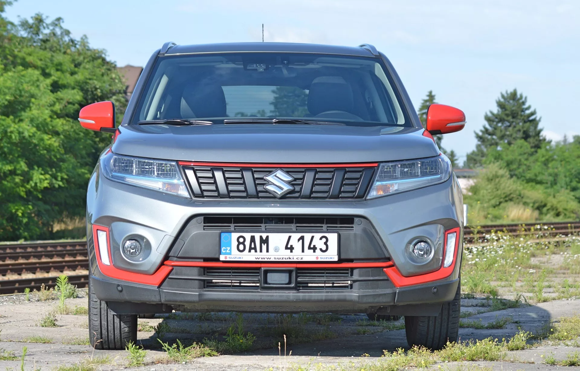 Přední pohled na vůz Suzuki Vitara 2018 s mírně nízkým úhlem. Obrázek zdůrazňuje přední rysy vozidla, včetně kapoty, mřížky (se znakem Suzuki), elegantních světlometů (potenciálně LED) a osvětlení nárazníků. Vůz vypadá robustně, což naznačuje schopnost jízdy v terénu. Viditelné jsou ukazatele směru v zrcátkách a přední poznávací značka. Šedá karoserie s červenými akcenty může znamenat speciální výbavu. Pozadí se jeví jako opuštěná průmyslová oblast nebo železniční dvůr, což poskytuje jasný pohled na design vozu.