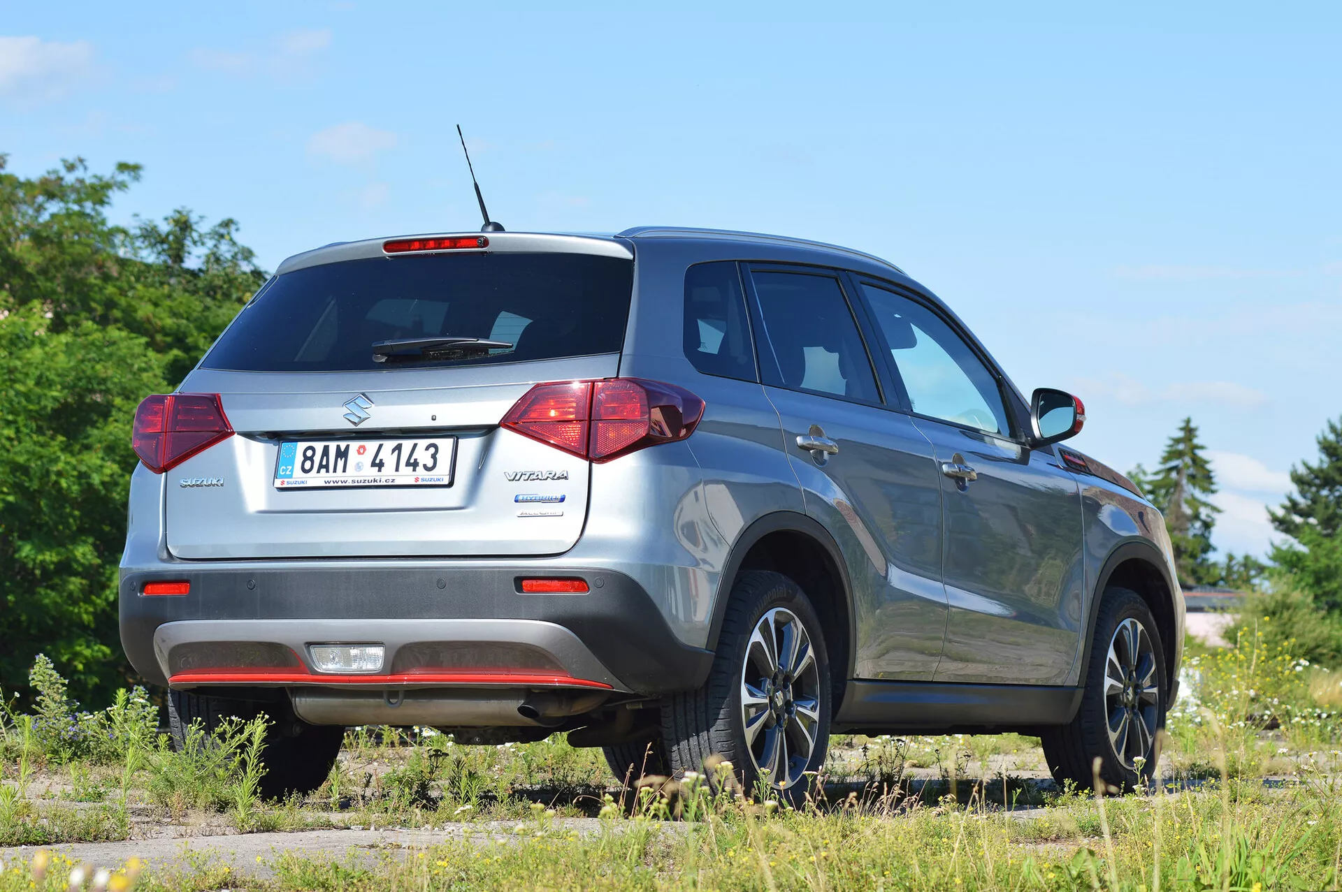 Suzuki Vitara 2018 zachycená z pravého zadního tříčtvrtečního úhlu, zdůrazňující design zadních světel, zadní okno a profil na straně spolujezdce. Obrázek zobrazuje moderní vybavení kompaktního SUV, jako je střešní anténa pro technologické funkce, zadní parkovací senzory a stylová litá kola. Vozidlo je umístěno na nezpevněném povrchu v venkovském prostředí, což naznačuje jeho schopnost off-road. Ideální pro kategorie automotive, SUV a obrázky tvořící off-road vozidla.