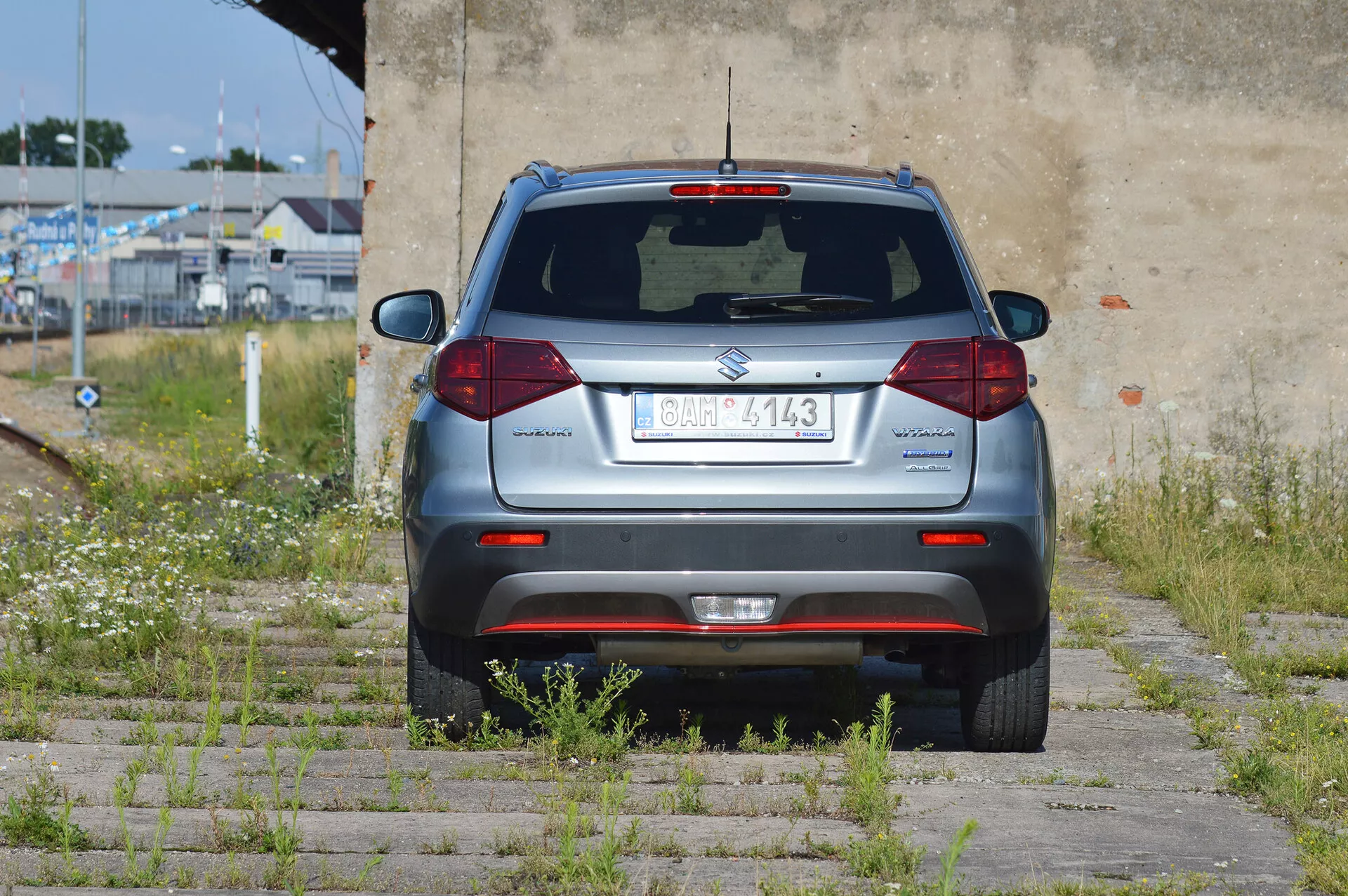 Zadní úhel stříbrného kompaktního SUV Suzuki Vitara 2018 zaparkovaného na trávě a betonovém dlažbě. Celá zadní část je viditelná, zahrnuje středovou poznávací značku, zadní stěrač, třetí brzdové světlo, standardní halogenová zadní světla, odrazky na nárazníku, parkovací senzory a výfukovou trubku vlevo. Vozidlo se nachází v poloopuštěné oblasti s přerostlou vegetací a zvětralou zdí.