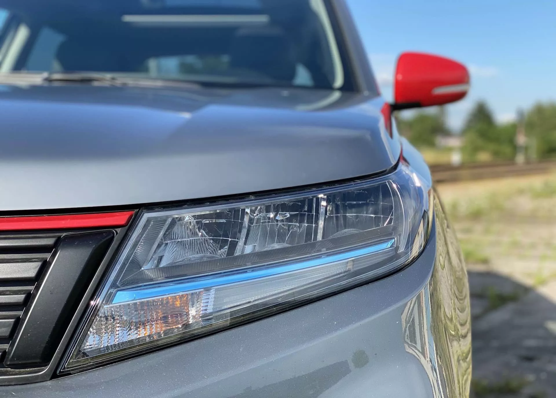 Detailní záběr na přední levý roh vozu Suzuki Vitara 2018 se zaměřením na LED světlomety s denními svítilnami, částečně viditelnou mřížku s vertikálními pruhy, jemně se svažující kapotu, kovový lak, kontrastní červené zpětné zrcátko pravděpodobně s integrovanými směrovými světly a rozmazané pozadí, které zdůrazňuje elegantní design a aerodynamiku.