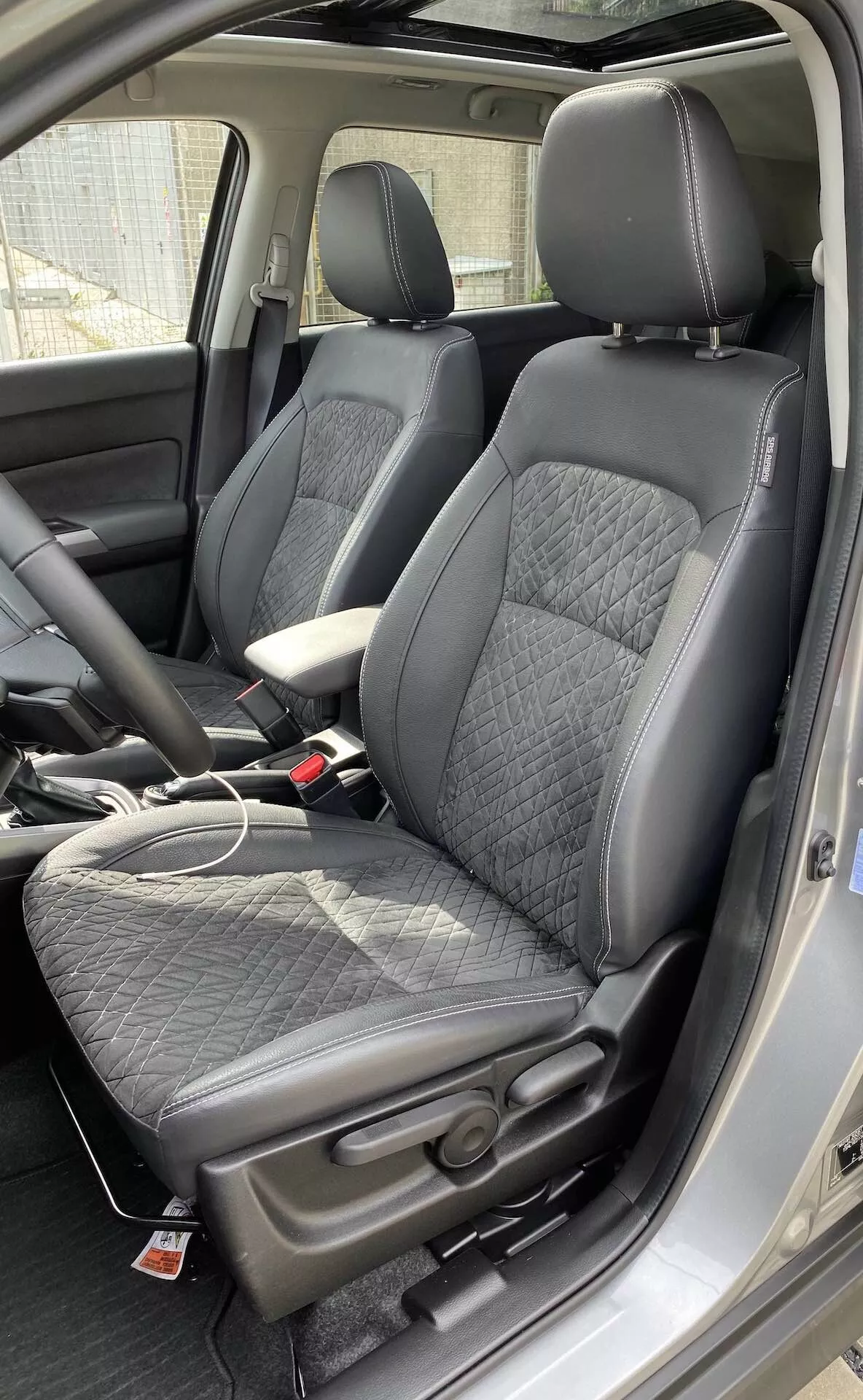 Interiér Suzuki Vitara 2018, zdůrazňující moderní přední sedadla ve stylu kyblíku z mírně nadhledové perspektivy. Sedadla jsou čalouněna černou kůží s prošívaným vzorem ve tvaru diamantů a boky s hladkou kůží, což nabízí rovnováhu mezi pohodlím a sportovností. Červený štítek na sedadlech naznačuje další funkce, jako jsou boční airbagy nebo vyhřívaná sedadla. Viditelná je také středová konzola s koženou opěrkou ruky, stejně jako nabíjecí kabel na sedadle spolujezdce, což naznačuje přítomnost nabíjecích portů. Zapadání bezpečnostních pásů odpovídá černému interiéru. Viditelná je také část přístrojové desky a B-sloupek, což naznačuje prostornou kabinu.