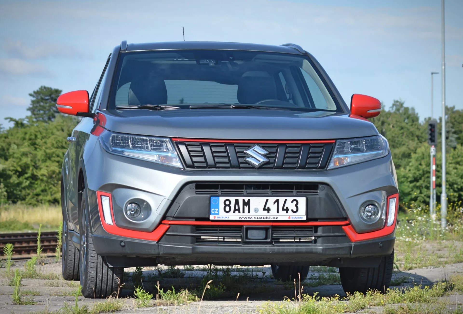 Obrázek ukazuje moderní SUV Suzuki Vitara 2018 z mírného předního úhlu, zvýrazňující přední fasci a část strany spolujezdce na pozadí řídké krajiny s železničními tratěmi. Auto, zaparkované na povrchu, který vypadá jako štěrkový, má výraznou mřížku s úhlovým designem, elegantní světlomety pravděpodobně s LED nebo HID technologií a integrované denní svícení nebo mlhovky v předním nárazníku. Nárazník zahrnuje kontrastní barevnou vložku, což naznačuje terénní nebo sportovní úroveň výbavy, a viditelná světlá výška zdůrazňuje charakter SUV. Zvýšené linie na kapotě přispívají k agresivnímu stylu, zatímco boční zrcátka a blatníky ukazují moderní design. Částečně viditelná litá kola doplňují robustní vzhled. Fotografie pořízená z blízkosti umožňuje podrobnou kontrolu těchto rysů.