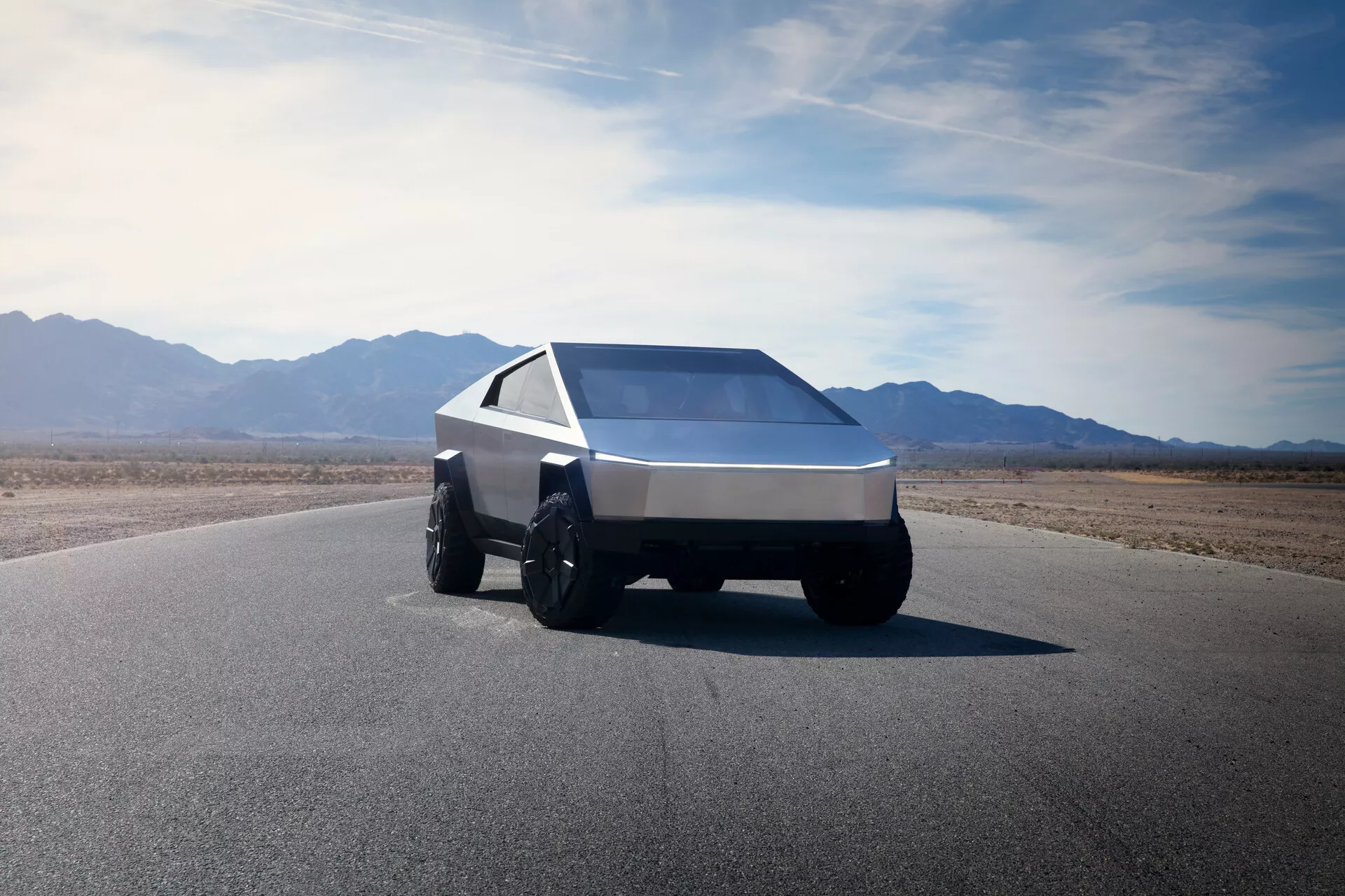 Tesla Cybertruck 2021 (1. generace) vyfotografovaná z nízkého předního tříčtvrtečního úhlu, předvádí svůj futuristický, úhlový design. Tento pohled zachycuje levý přední bok vozu, zdůrazňující klínovitý tvar s ostrými liniemi a rovnými plochami. Vozidlo je vybaveno velkými, robustními pneumatikami vhodnými pro jízdu v terénu a obsahuje jediné, nepřerušené světelné pásmo přes šířku přední strany, které naznačuje moderní LED nebo laserové osvětlení. Strmě skloněné, rozměrné čelní sklo a ostře zužující se boční okna přispívají k jeho geometrickému vzhledu. Vůz je zhotoven z kartáčovaného kovu nebo kompozitního materiálu, což naznačuje trvanlivost a robustnost. Pozadí obsahuje rozsáhlé čisté nebe a hornatý terén, naznačující suché, pouštní prostředí.