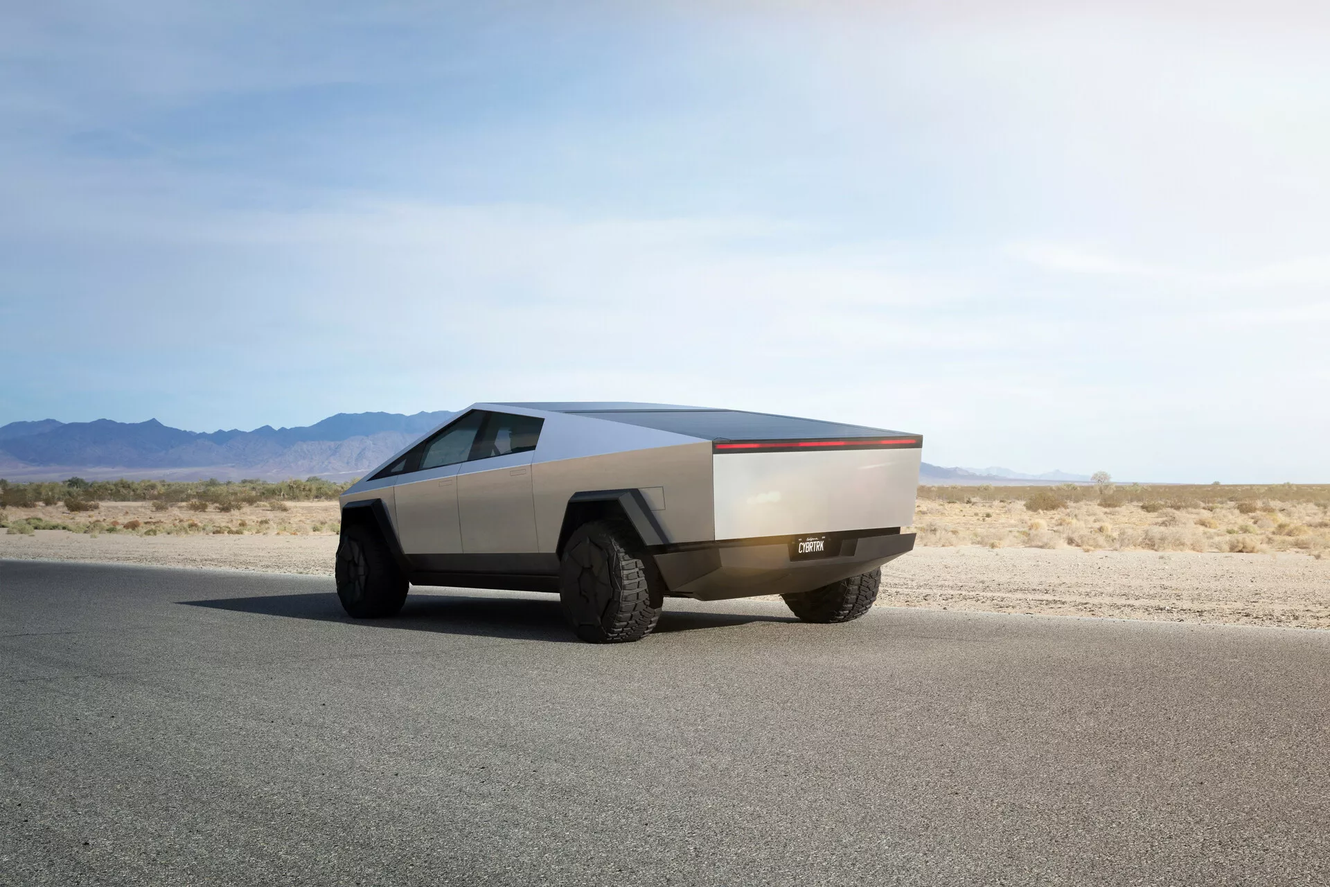 Futuristický Tesla Cybertruck 2021 na rovném asfaltovém povrchu zachycený z úhlu třech čtvrtin zezadu, ukazuje jeho charakteristický hranatý design, kovovou povrchovou úpravu, velké terénní pneumatiky a absence tradičních zrcátek a klik dveří, což naznačuje pokročilé automatizované mechanismy.