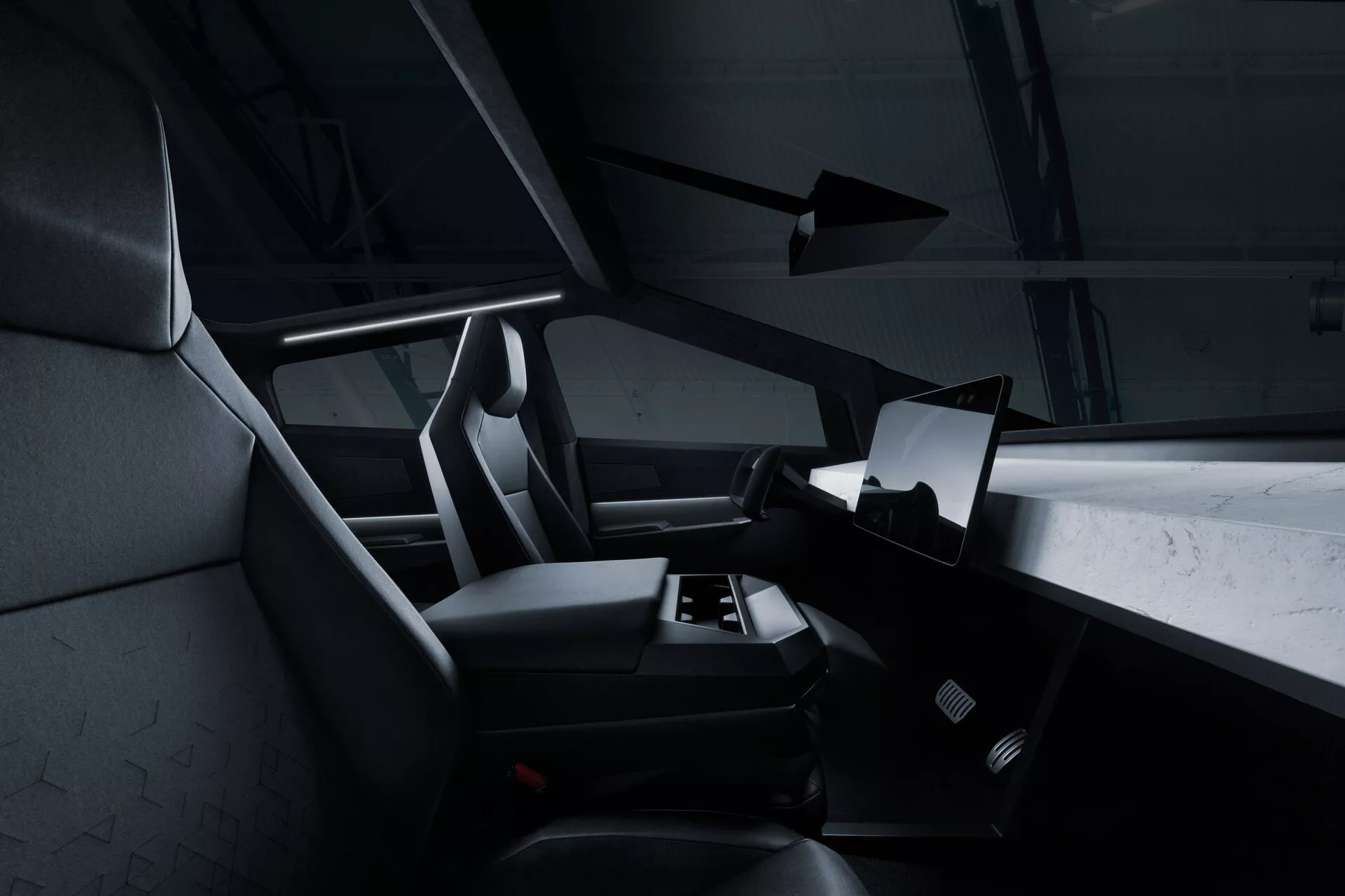 Pohled na interiér Tesla Cybertruck 2021 (1. generace) ze zadního sedadla, zobrazující pravou stranu kabiny s velkou horizontálně orientovanou dotykovou obrazovkou uprostřed palubní desky. Částečně je vidět volant, ale je většinou zakrytý. Přední sedadla a sedadlo řidiče mají integrované opěrky hlavy a texturované čalounění, pravděpodobně z tkaniny nebo syntetické kůže. V oblasti pod nohama je patrná autokoberce a nejsou viditelné žádné manuální ovládací prvky, což naznačuje minimalistický, digitální přístup. Tmavá barevná paleta a jemné akcentní osvětlení dodávají moderní, luxusní vzhled.