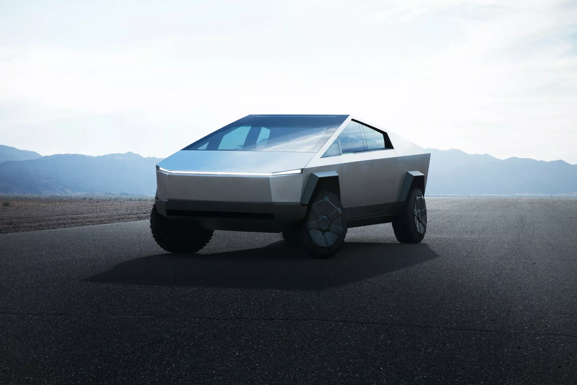 Obrázek vozu Tesla Cybertruck 2021 (1. generace) na silnici s plochým terénem nebo pouštními horami v pozadí za denního světla a jasné oblohy. Fotografie je pořízena z nízkého úhlu, čímž poskytuje záběr na přední čtvrtinu vozu a zdůrazňuje futuristický, hranatý design vozidla. Vozidlo má klinovitý předek, tělo z plochých panelů, tenké horizontální světlomety, kontinuálně svažitý čelní sklo, žádné viditelné boční zrcátka, velká kola schopná terénního provozu s propracovanými ráfky a postrádá přední mřížku, což naznačuje elektrický pohon.