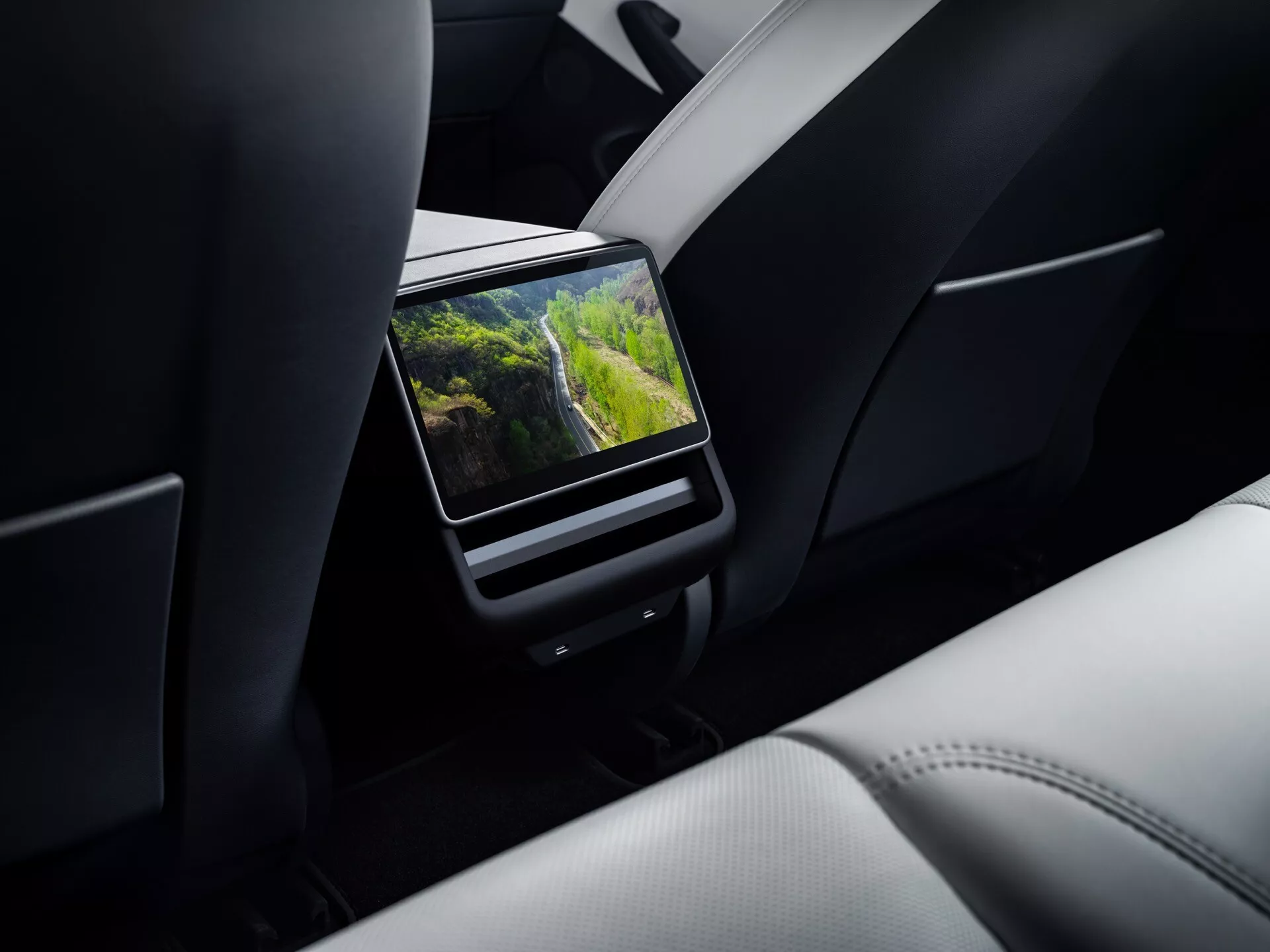 Tesla Model 3 (1. generace facelift) rok 2023 prezentující detailní pohled na OEM zábavní systém na zadním sedadle s vysokým rozlišením mezi sedadlem spolujezdce a řidiče, zobrazující živý venkovní scénář na displeji v luxusní kabině s prvotřídním koženým čalouněním.