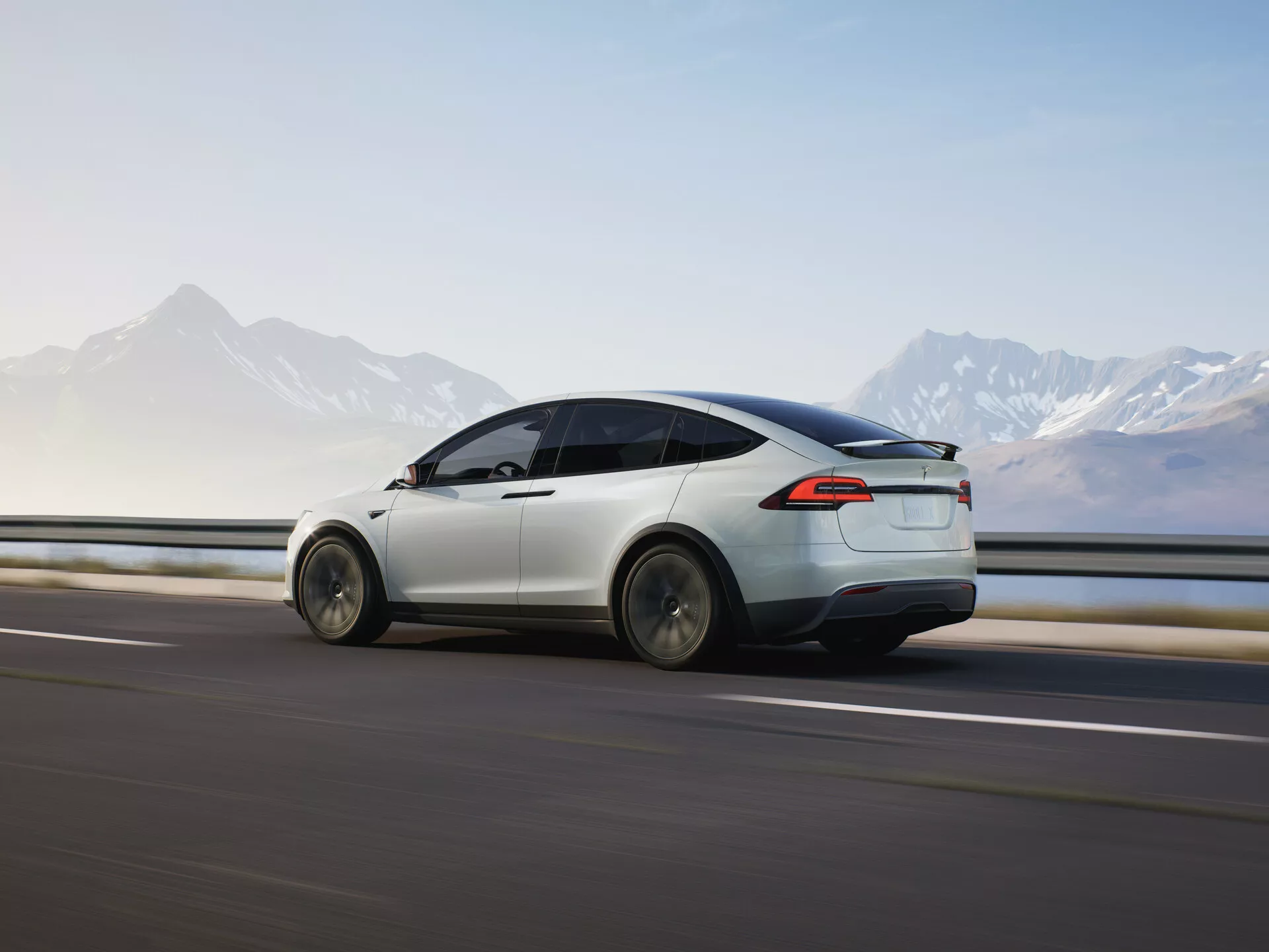 Dynamický pohled na zadní třetinu 2021 Tesla Model X (1. generace-facelift) v pohybu, zvýrazňující jeho aerodynamický design, elegantní bílý lak, LED zadní světla a moderní design kol. Auto je zachyceno při pohybu zleva doprava proti rozmazané silnici a svodidlu, což naznačuje rychlost, s horským horizontem v pozadí. Měkké osvětlení naznačuje časné ráno nebo pozdní odpoledne, což zdůrazňuje čisté elektrické linie a pokročilé funkce vozidla v malebném prostředí.