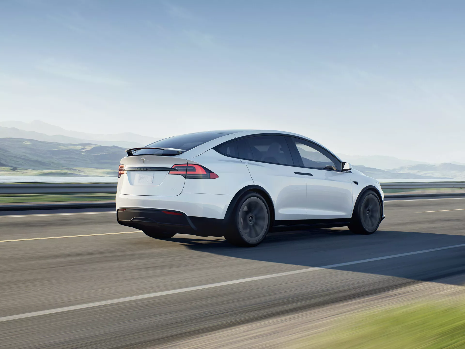 Dynamický obrázek bílého elektrického hatchbacku sedan Tesla Model X 2021 v pohybu, zobrazený ze tříčtvrtečního úhlu zezadu. Fotografie zachycuje levý bok vozidla z mírně zvýšené perspektivy, zdůrazňuje zadní kola, koncová světla a konturu zadní části vozu, s důrazem na jeho aerodynamický design se skloněnou střechou a hladkými křivkami. Design naznačuje minimalizované poruchy vzduchu a nízký koeficient odporu, podpořený koly navrženými pro aero efektivitu. Zadní okno přechází do černé skleněné střechy, což naznačuje panoramatické střešní okno, které symbolizuje moderní automobilovou technologii. Elegantní LED koncová světla jsou výraznou charakteristikou současné osvětlovací technologie. Absence výfukové trubky indikuje plně elektrickou povahu vozu. Vhodné pro kategorie jako vozidla, elektrická vozidla, pohybová fotografie, automobilový design, moderní technologie a luxusní vozidla.