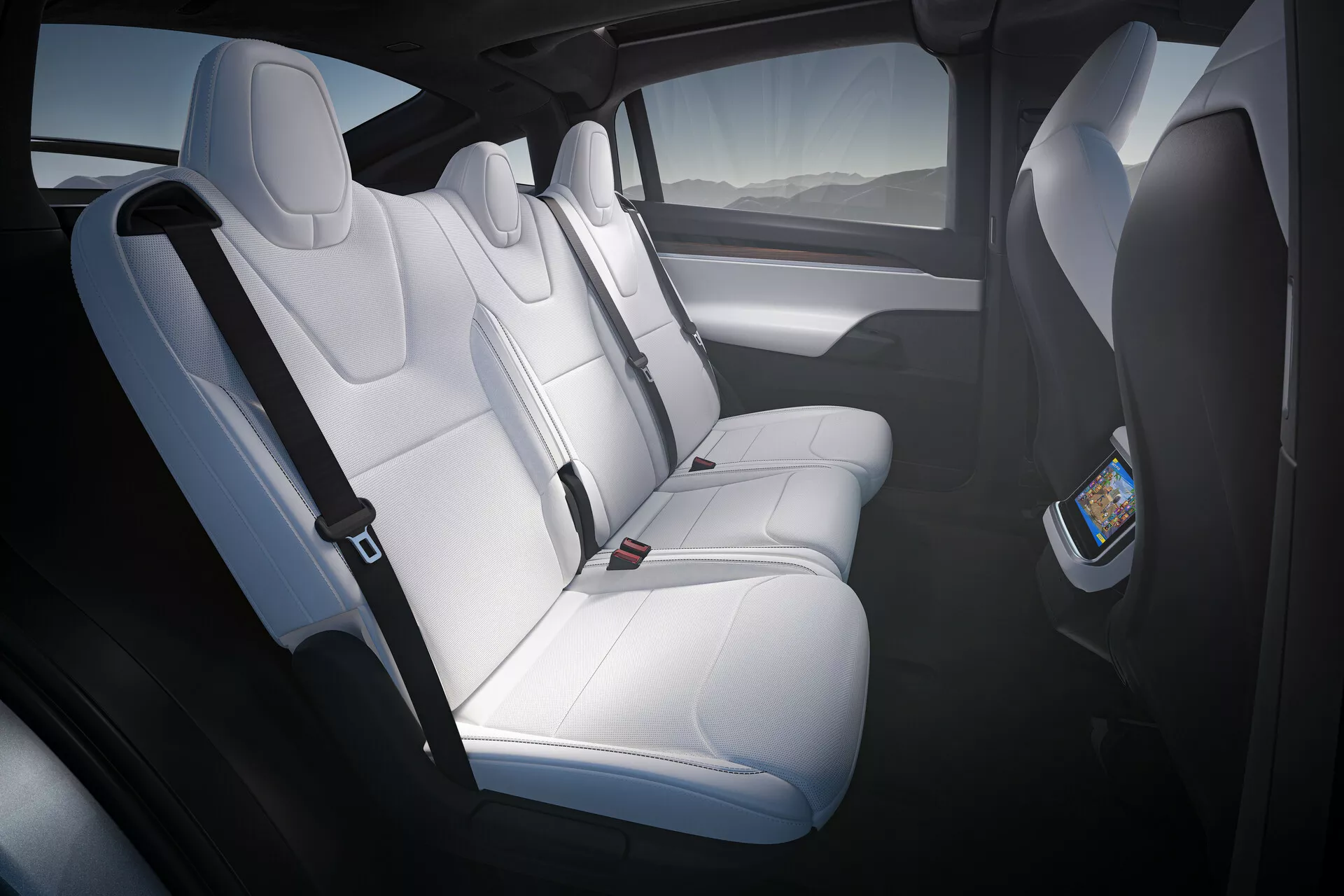 Interiérový záběr na Tesla Model X 2021 (1. generace-facelift), zobrazující zadní prostor pro cestující ze zadních dveří na straně řidiče. Bílé kožené sedadla s opěrkami hlavy a tvarovaným polstrováním pojmou tři pasažéry. Digitální displej na zadní straně sedadla předního spolujezdce naznačuje systém zadní zábavy. Prostorná kabina, možná s panoramatickým střešním oknem, používá přirozené světlo k zvýraznění kontrastních tmavých dveřních panelů s integrovanými mřížkami reproduktorů.