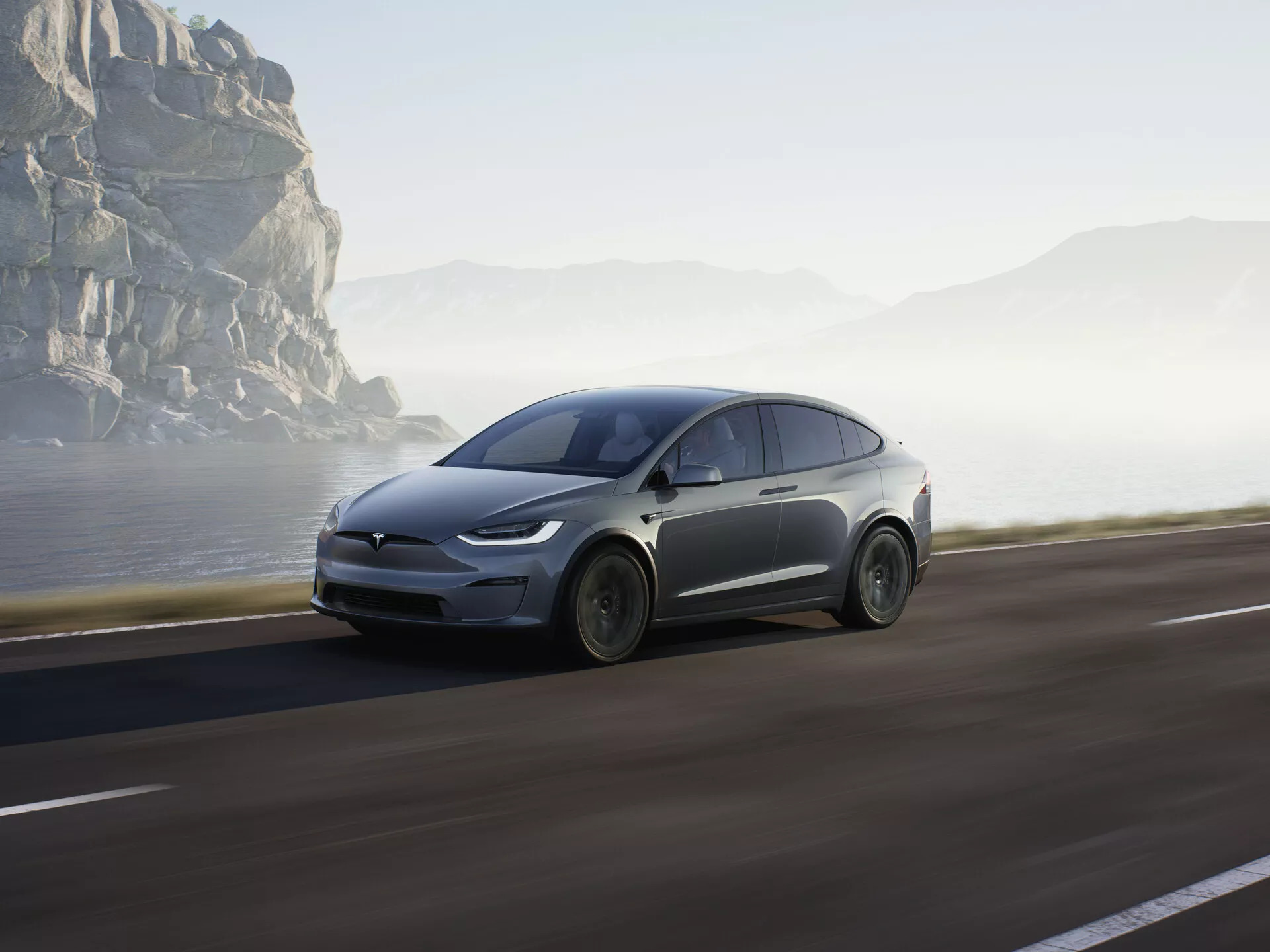 Obrázek ukazuje moderní elektrické SUV, konkrétně Tesla Model X 2021 (1. generace facelift), jedoucí po pobřežní silnici. Je zachyceno z nízkého bočního profilu, který zvýrazňuje přední čtvrtinu s důrazem na přední kolo, kapotu, čelní sklo a část dveří řidiče. Vozidlo zaujímá podstatnou část spodní poloviny rámu, což zdůrazňuje jeho dominanci. Auto je v pohybu s mírným rozmazáním kol a pozadí, což vytváří dojem rychlosti a dynamiky. Měkké sluneční světlo naznačuje časné ráno nebo pozdní odpoledne, což přidává scéně klidnou náladu v malebném a dobrodružném pobřežním prostředí. Lze předpokládat aerodynamické designové prvky a pokročilé funkce, jako jsou LED světla, což naznačuje pokročilou technologii jako regenerativní brzdění a možná i autonomní řízení. Auto je umístěno na pravé straně silnice a pohybuje se vpřed směrem k otevřenému cíli, přičemž pobřežní skály a mlha v pozadí zvýrazňují jeho futuristický vzhled a přirozené prostředí.