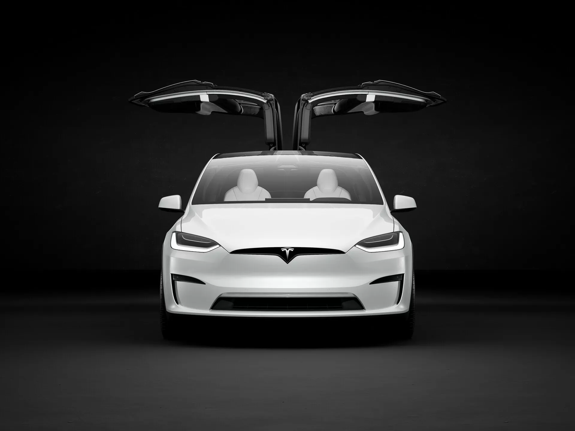 Obrázek představuje elektrické vozidlo Tesla Model X 2021 (první generace, facelift) v ateliérovém prostředí s tmavým pozadím, které kontrastuje s světlou barvou auta. Pohled je lehce z nízkého úhlu a zvýrazňuje přední část auta, včetně kapoty, nárazníku a oblasti mřížky, i když vzhledem k tomu, že se jedná o elektrické vozidlo, tradiční otevřenou mřížku nemá. Viditelný je také dolní přívod vzduchu. Obě přední dveře jsou otevřené, což odhaluje unikátní dveře typu falcon-wing, které se vyklápějí nahoru, což je typickým znakem luxusních modelů. Interiér ukazuje dvě řady světlých sedadel, což naznačuje minimalistický, ale luxusní design. Ostrá, hranatá LED světla dodávají agresivní nádech a logo Tesla je zřetelně umístěno nad oblasti přívodu vzduchu a pod předním kufrem - 'frunkem'. Auto naznačuje pokročilé technologie jako autonomní řízení, elektrický pohon, regenerační brzdové systémy, konektivitu a pokročilé bezpečnostní systémy, cílené na publikum, které ocení inovace, výkon a ekologické uvědomění.