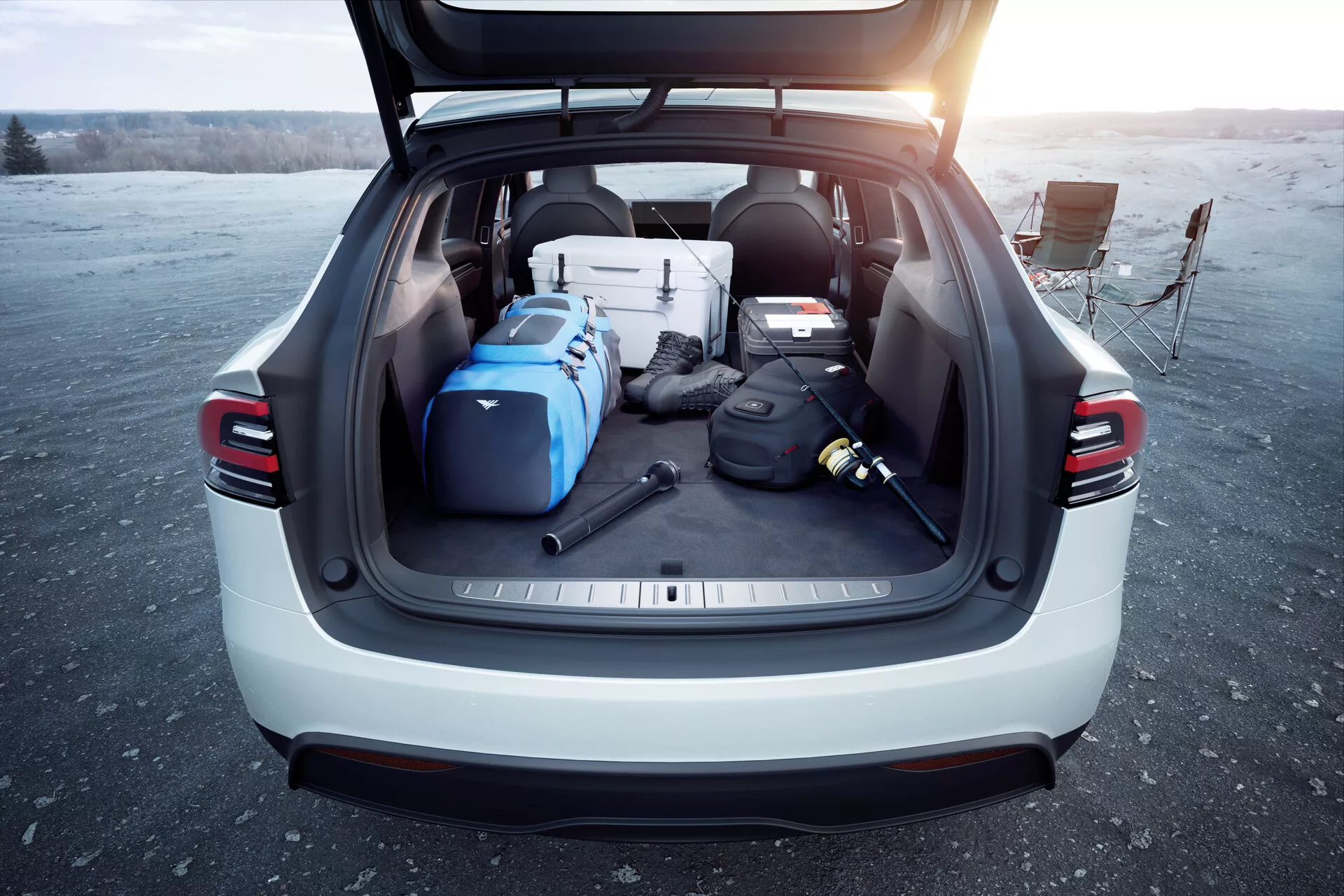 Zadní pohled na SUV Tesla Model X 2021 s otevřeným kufrem, ukazující prostorný nákladní prostor naplněný různými předměty za denního světla, pravděpodobně při východu nebo západu slunce. Obrázek zdůrazňuje užitečnost a moderní vlastnosti vozidla, jako jsou automatická zadní vrata, LED zadní světla, světlá barva exteriéru a kryt nákladu, foceno na otevřeném prostoru.
