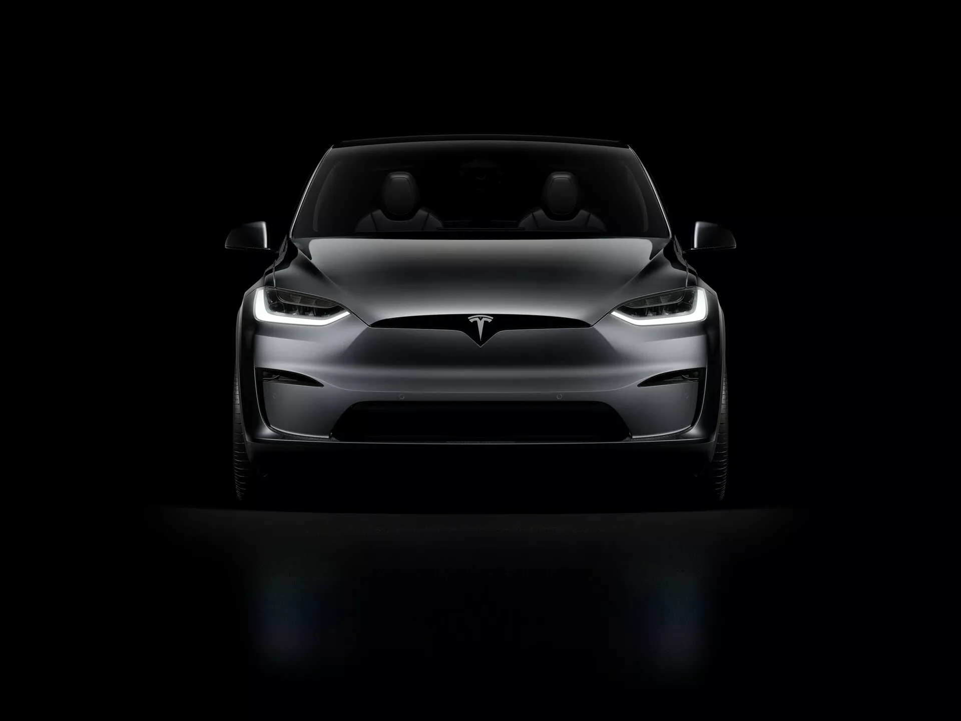Tesla Model X 2021 (1. generace facelift) fotografovaná z předního pohledu v nízkém úhlu. Obraz zdůrazňuje přední část vozu, LED světlomety, elegantní návrh kapoty, hladkou přední část bez mřížky, minimalistický přední nárazník a aerodynamické boční zrcátka. Dramatické osvětlení zvýrazňuje hlavní prvky, zatímco vůz stojí na černém pozadí, což zvyšuje jeho impozantní postoj.