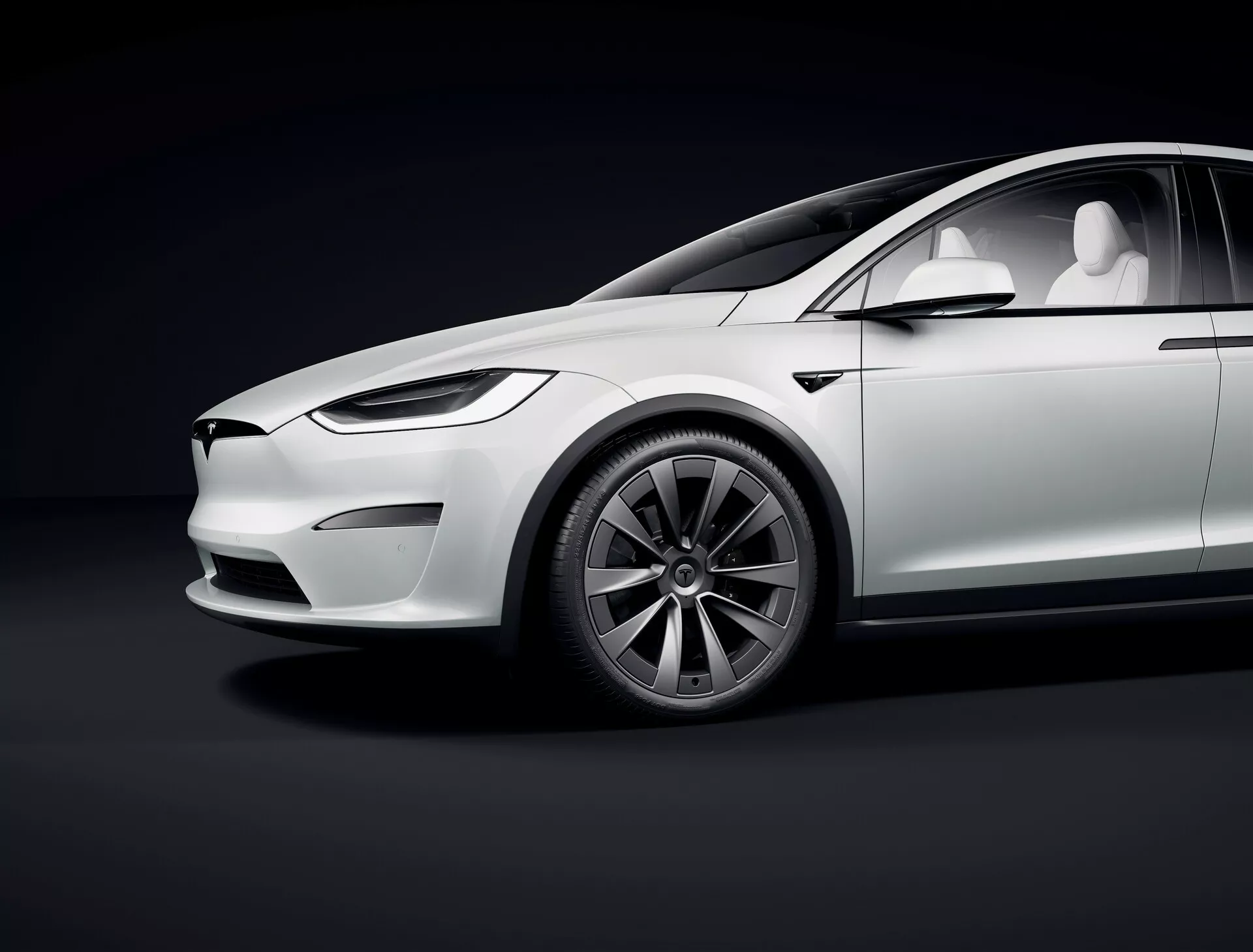 Studiový snímek představující boční profil Tesla Model X 2021 první generace napříč změnami, z nízkého úhlu, zdůrazňující luxusní elektrické prvky, jako je elegantní design, absence výfukových trubek, minimalistická přední část s LED světlomety, zatahovací kliky dveří, vícepaprsková aero kola, velké nízkoprofilové pneumatiky, bílý lesklý lak s jemnými odrazy, panoramatické střešní okno a kamery místo zpětných zrcátek. Křivky a linie vozu jsou zvýrazněny studiovým osvětlením proti tmavému gradientnímu pozadí.