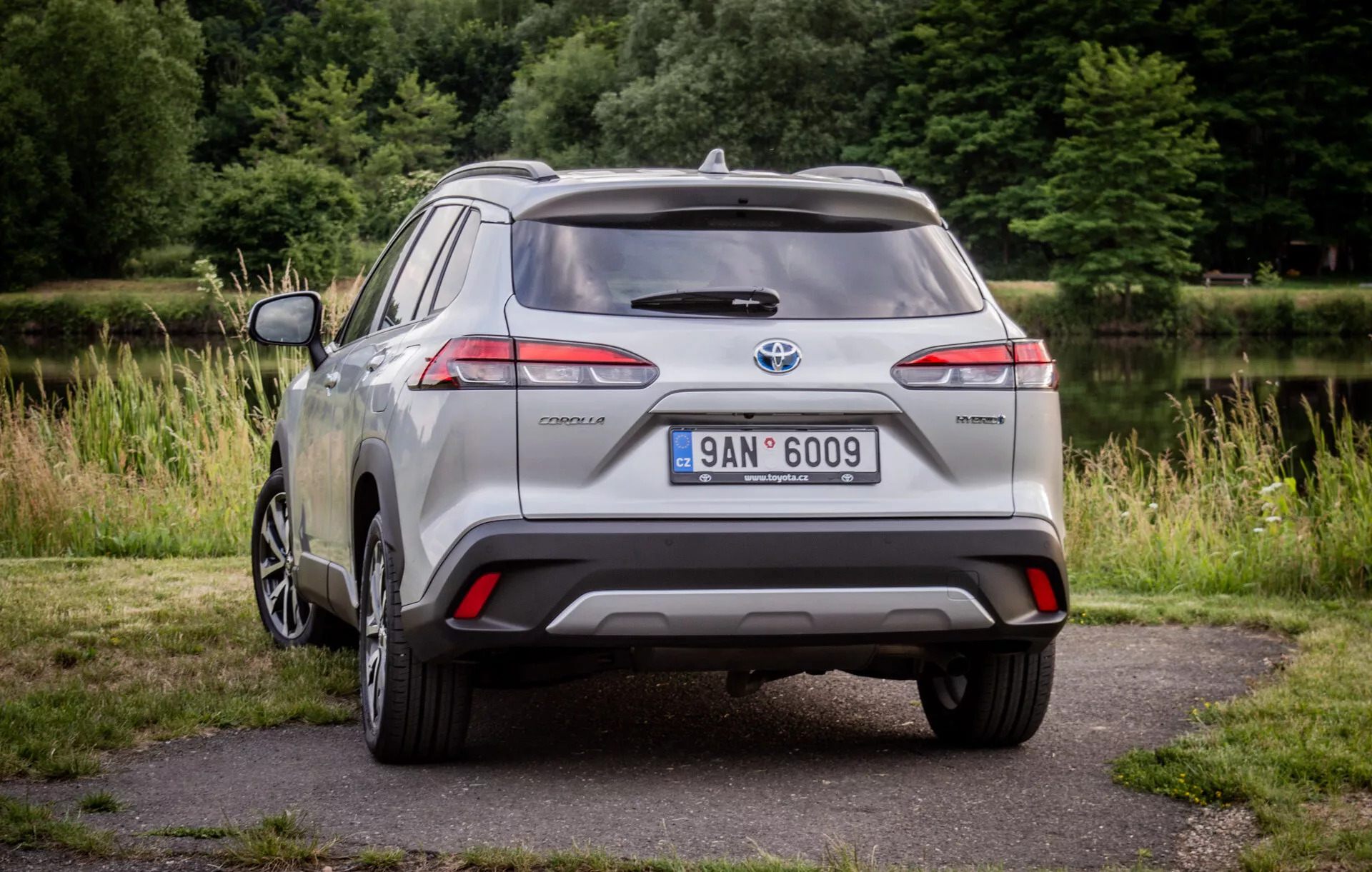 Stříbrný Toyota Corolla Cross z roku 2022 zaparkovaný na dlažbě se zelení v pozadí ukazuje jeho LED zadní světla, zadní spoiler, slitinové kolo s více paprsky a označení hybrid v přirozeném denním světle
