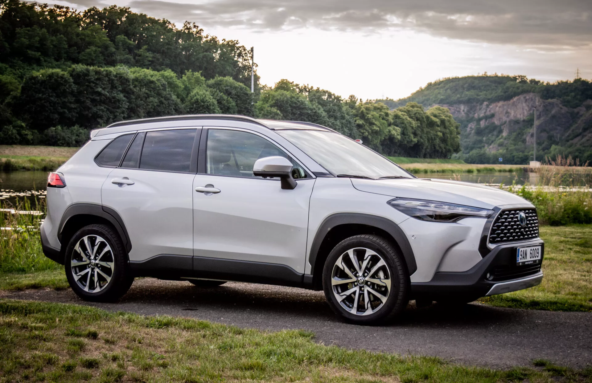 SUV Toyota Corolla Cross 2022 zaparkované venku, předvádějící svůj moderní design z čelně-bočního úhlu s důrazem na mřížku chladiče, slitinová kola a boční profil na pozadí s přírodní scenérií