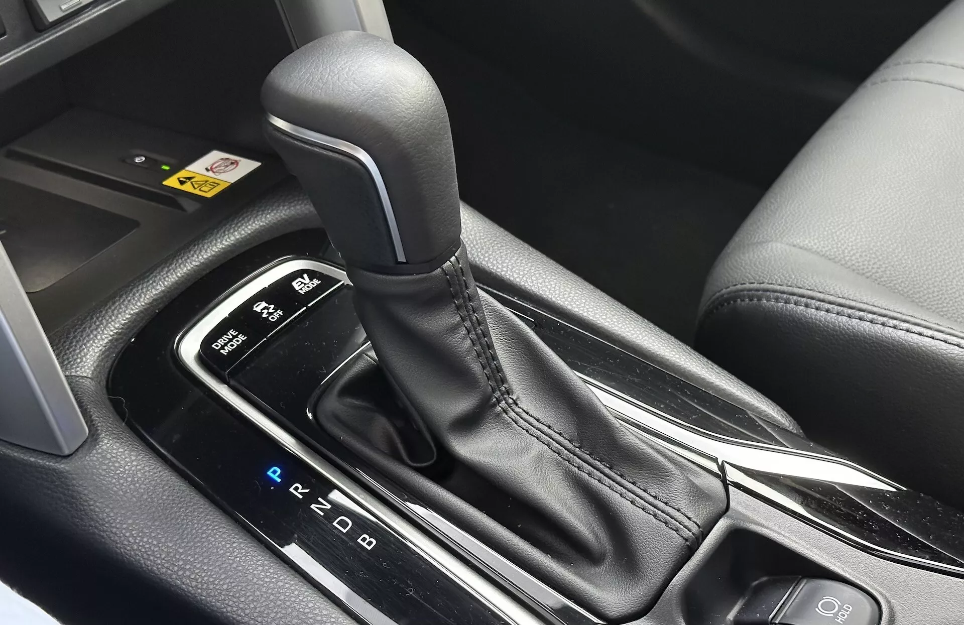 Detailní interiérový pohled na Toyota Corolla Cross 2022 ukazující automatickou řadicí páku s viditelným rozložením PRND, obklopenou elegantní středovou konzolou s černými plastovými a kovovými prvky, zdůrazňující ergonomický design a moderní technologické funkce