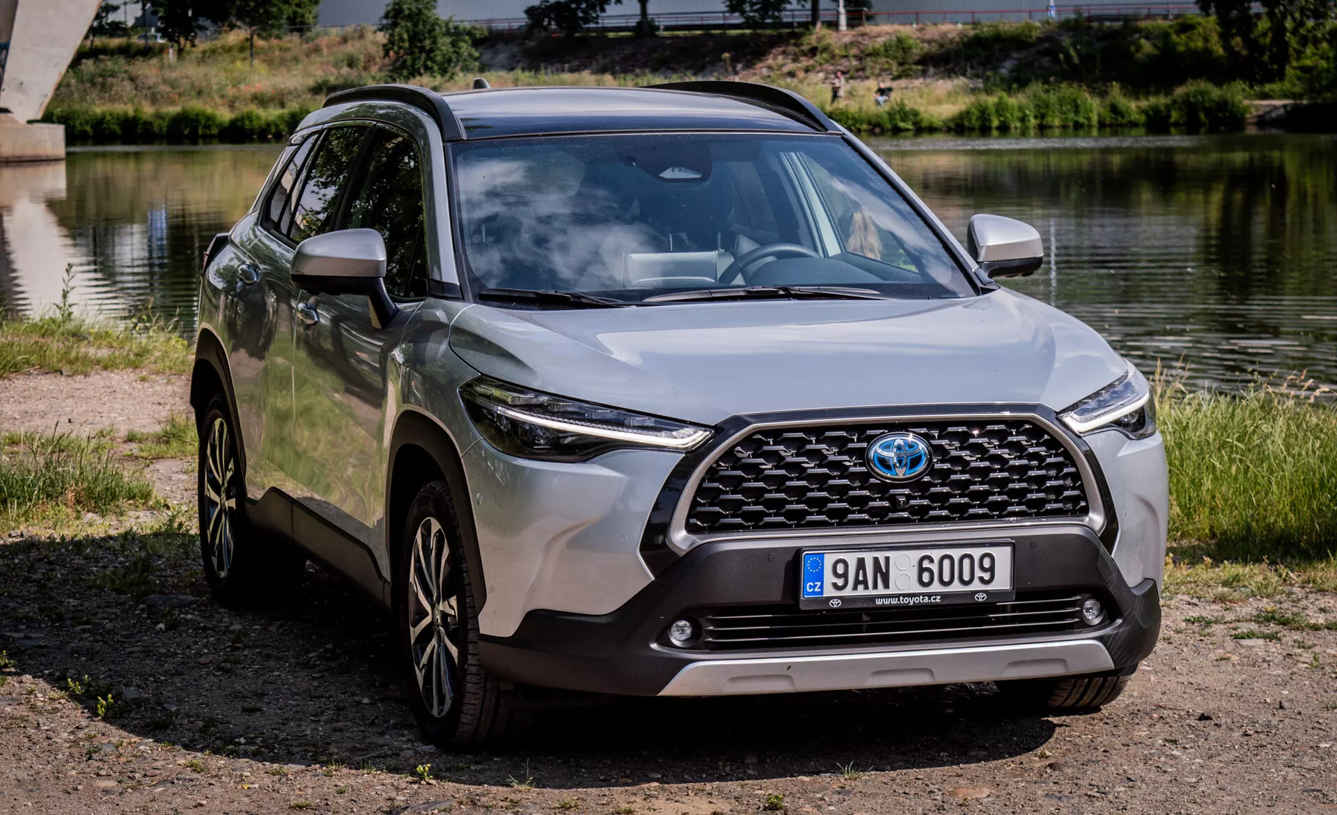 Moderní crossover SUV Toyota Corolla Cross 2022 zaparkované venku poblíž vodní plochy s čistým pohledem na jeho přední mřížku, kapotu, LED světlomety, přední nárazník a profil strany včetně slitinových kol a karoserií lakovaných bočních zrcátek