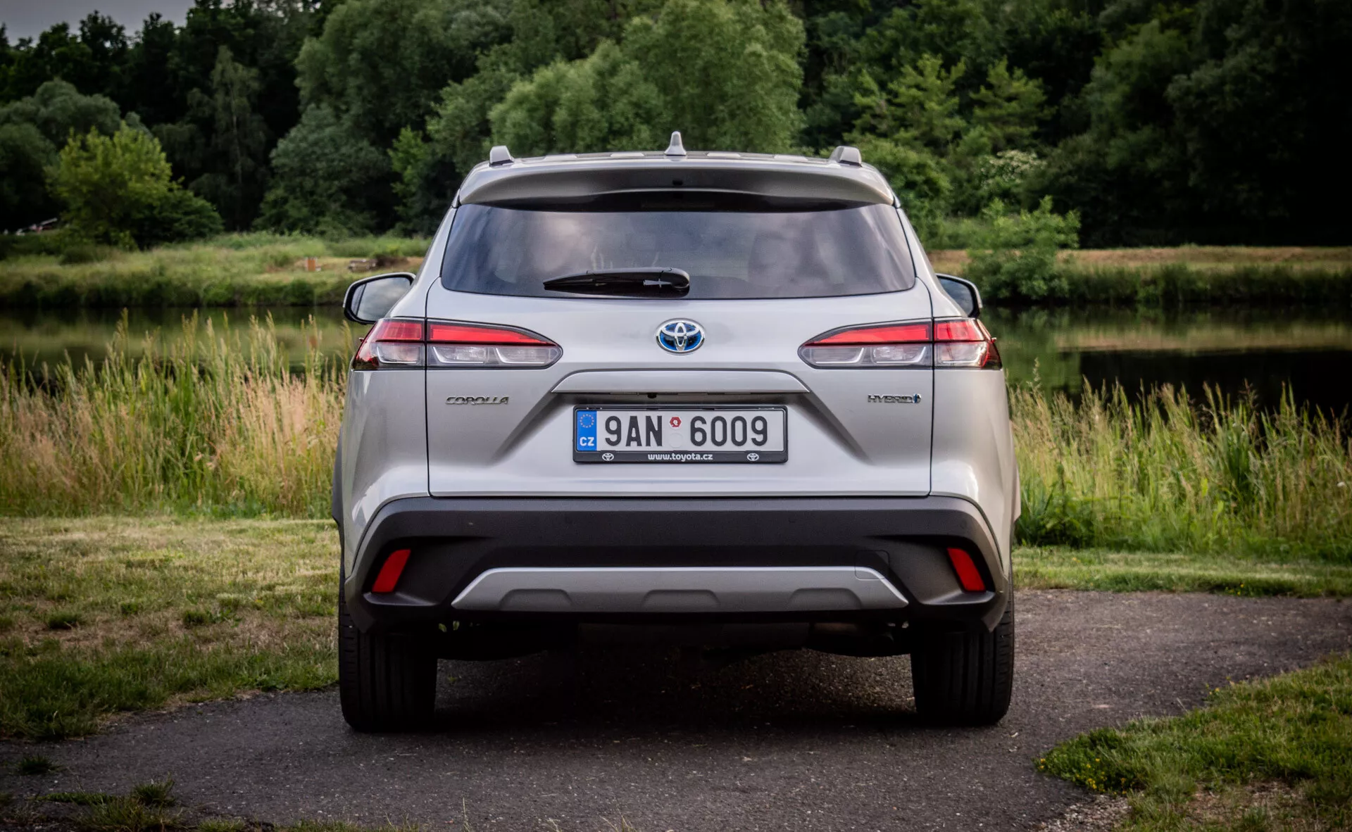 Zadní pohled na hybridní model Toyota C-HR s LED zadními světly, integrovaným difuzorem nárazníku se světly, anténou na střeše a slitinovými koly, umístěným na přírodním pozadí