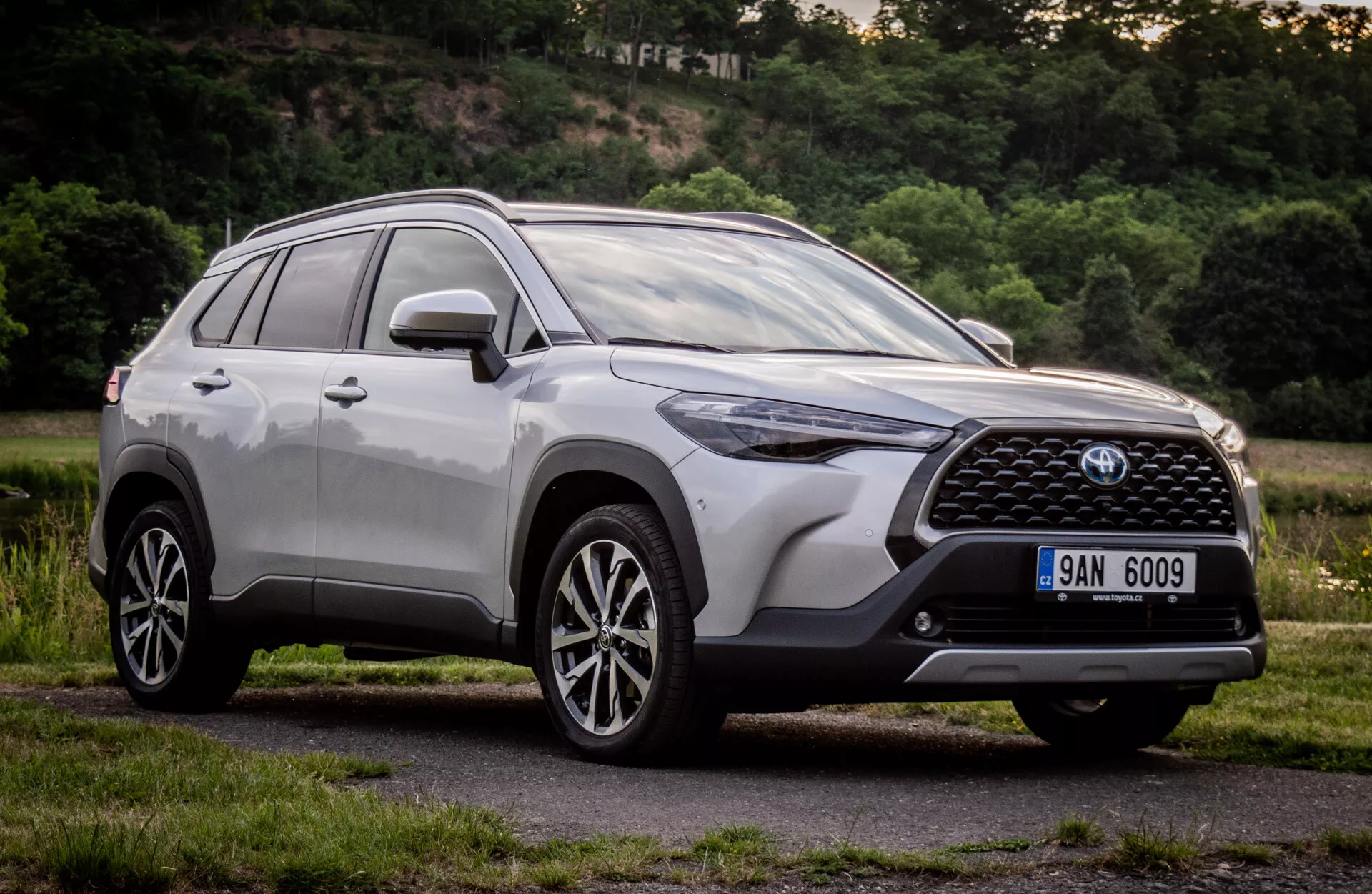 Toyota Corolla Cross 2022 v lesklém kovovém provedení ukazuje svůj moderní design SUV s detailním předním grilem, emblémem, úhlovými LED světlomety, aerodynamickým předním nárazníkem, konturovanou kapotou, bočním profilem s litými koly s více paprsky a střešními ližinami, snímek pořízený z nízkého úhlu k zdůraznění jeho výšky a postoje během dne v přírodním předměstském prostředí.